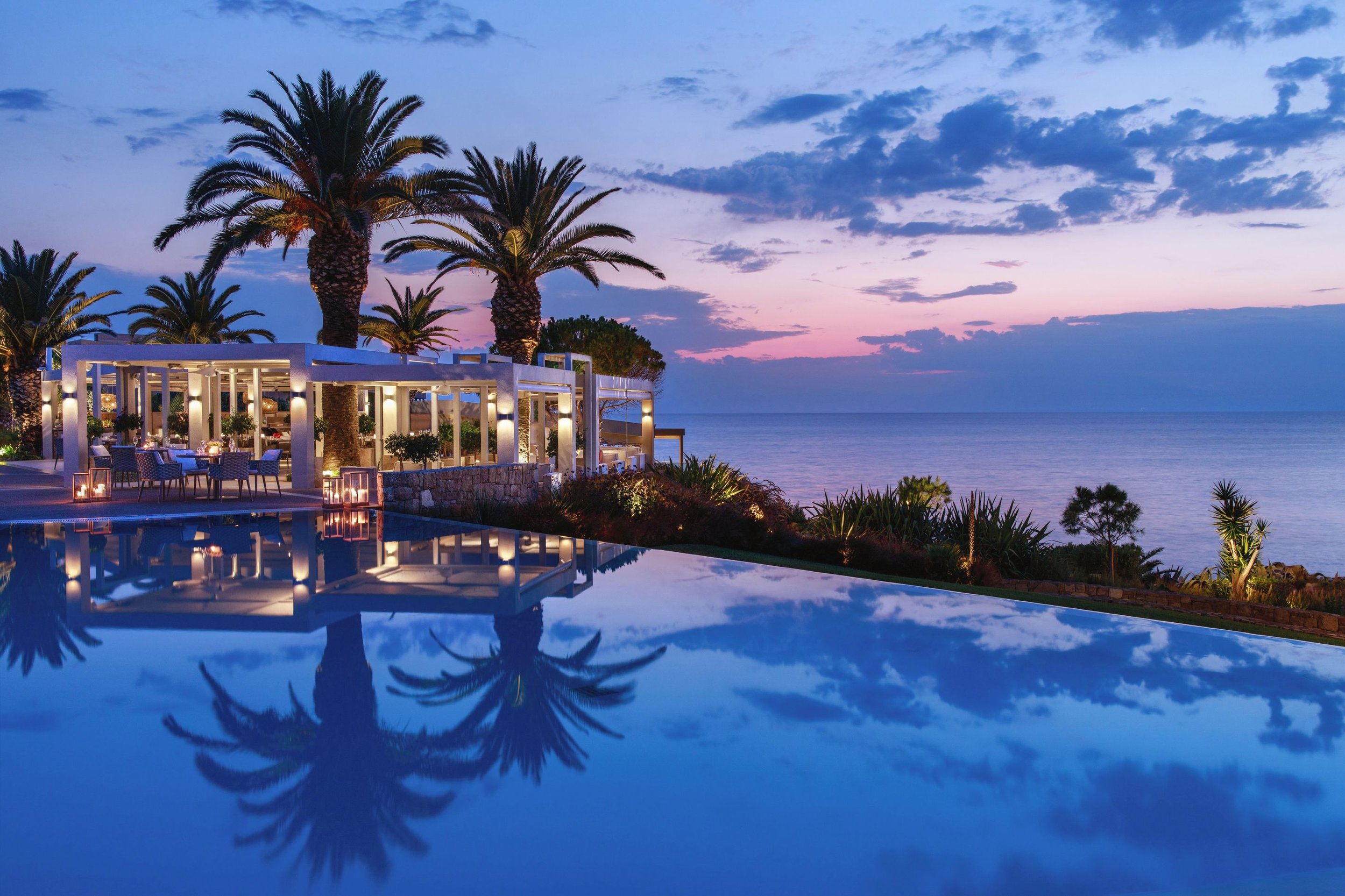 A luxurious outdoor pool reflects the evening sky with purple and blue clouds. Behind the pool, there are tall palm trees and a white modern cabana with seating and decorations illuminated by soft lighting, overlooking the ocean during sunset.