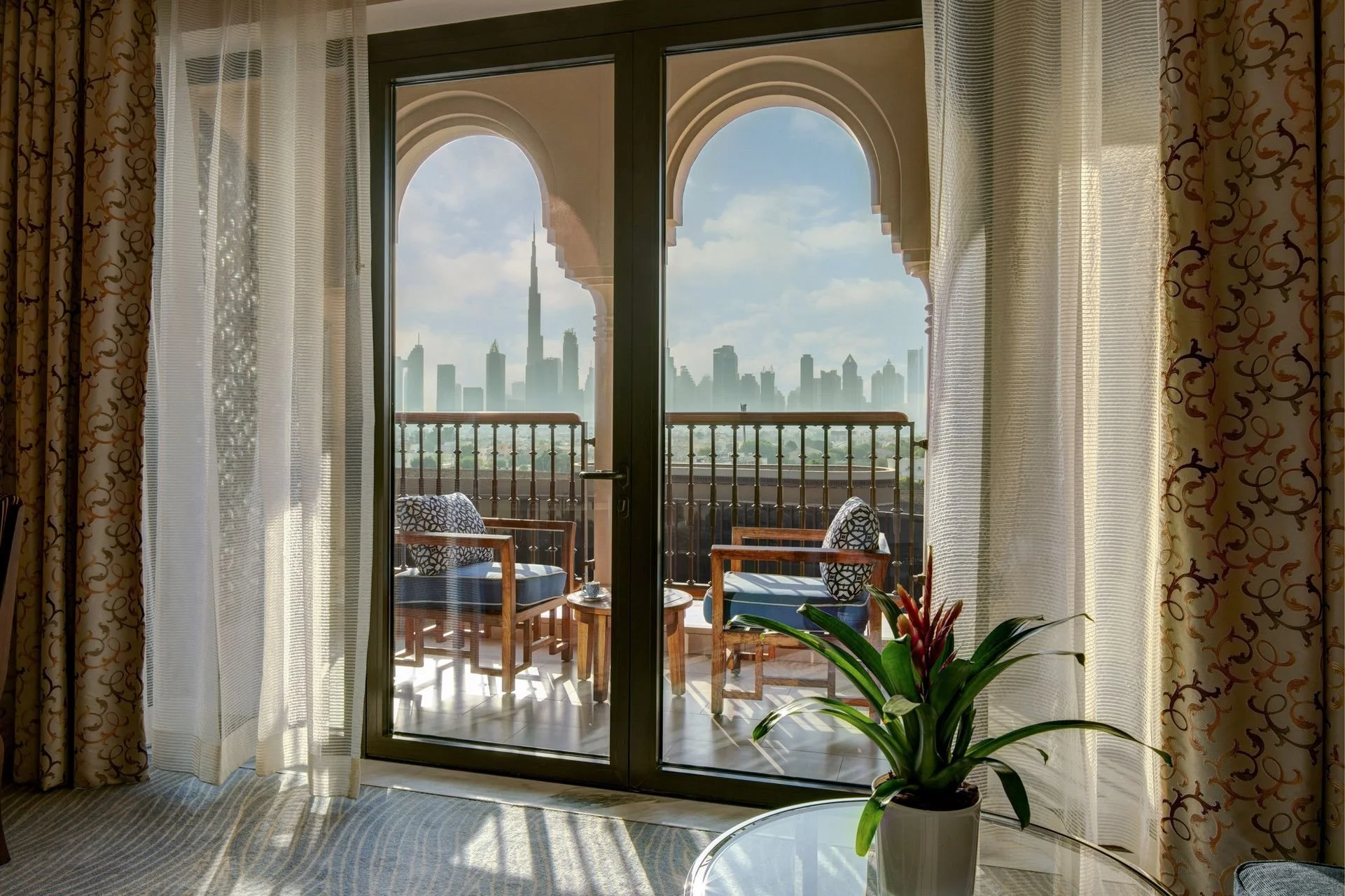 View of Dubai skyline with a balcony, framed by curtains, seen through glass doors, with a potted plant and a table inside.
