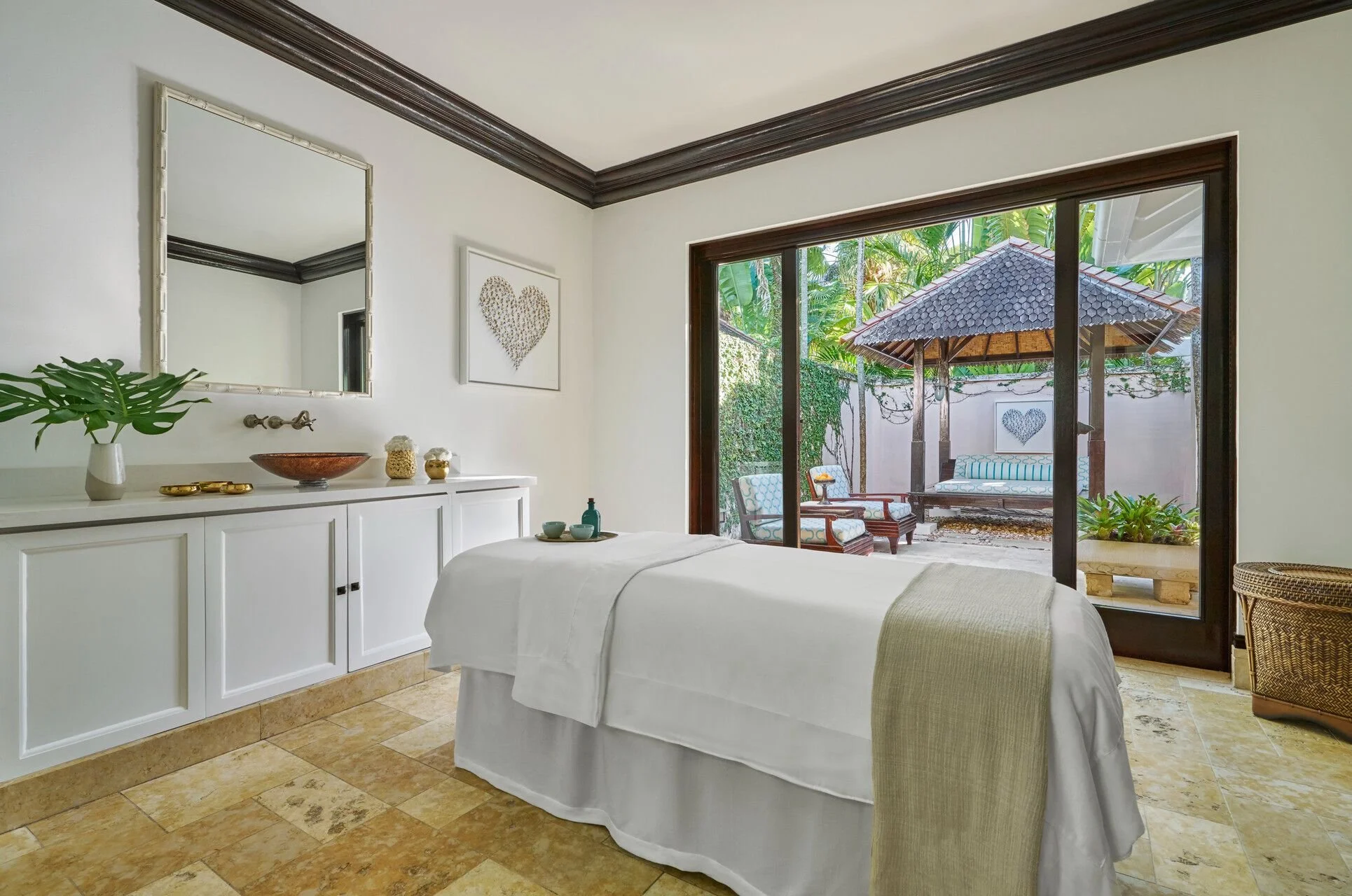 A bedroom with a bed covered in white linens and a beige throw. There is a white cabinet with decorative bowls, vases, and a plant on top. A large mirror hangs on the wall above the cabinet. Outside the sliding glass door, there is a patio area with outdoor seating, a small thatched roof gazebo, and lush green tropical plants.