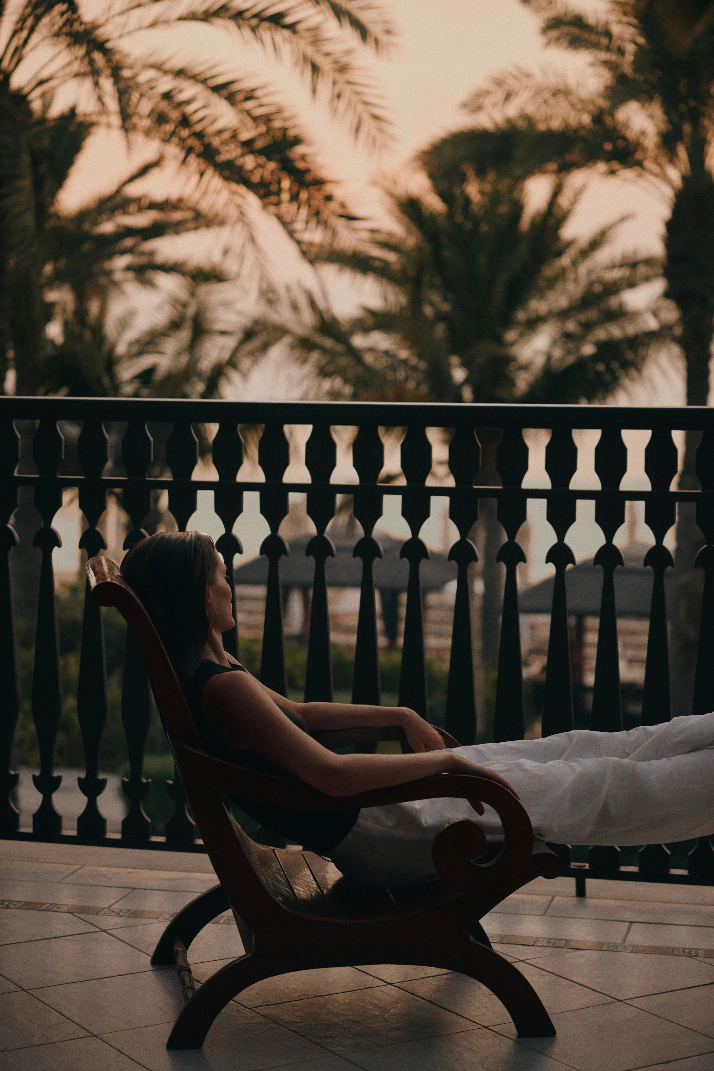 A woman sitting on a lounge chair with white sheets, relaxing on a balcony during sunset. Palm trees are visible in the background, with warm, golden light.