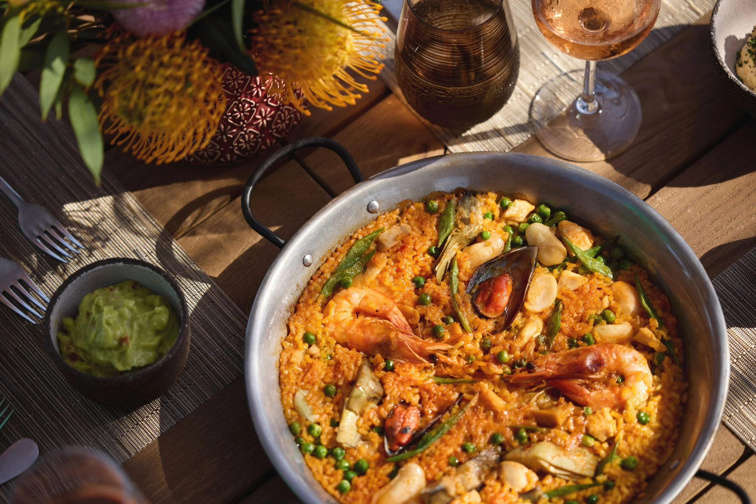A dish of seafood paella with shrimp, mussels, and peas in a large metal pan. Surrounding it are small bowls of guacamole and a bottle of dark soda. There are wine glasses, and a bouquet of flowers with yellow and purple blossoms on a wooden table.
