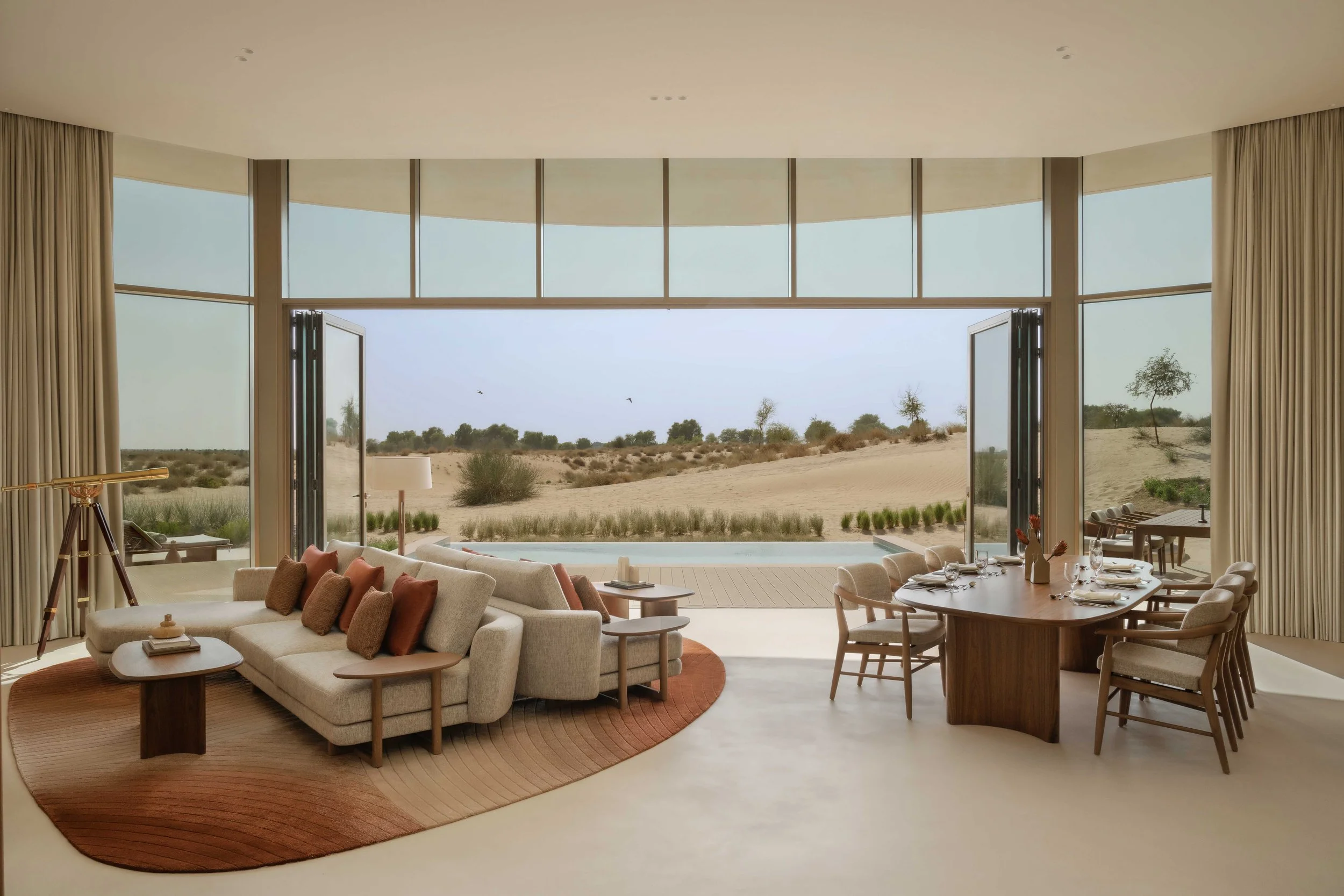Living room with large windows opening to desert landscape, furnished with beige sofa, armchairs, round wooden table, dining table set with chairs, and a telescope.