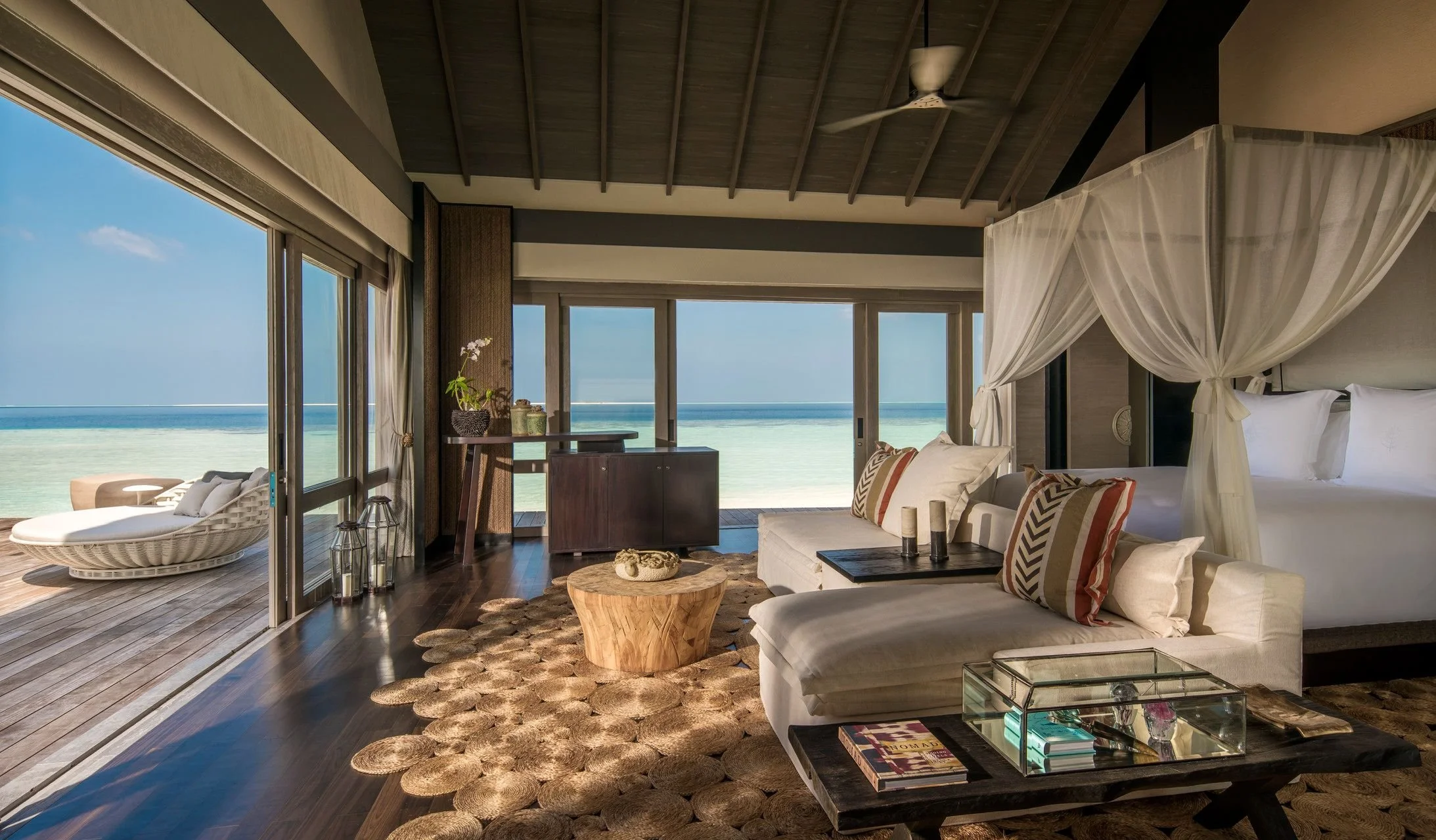 Interior of a luxurious beachside hotel room with large sliding glass doors open to the beach, featuring a white canopy bed, a sofa with patterned pillows, a wooden coffee table, and decorative items, with a view of the ocean and sand outside.