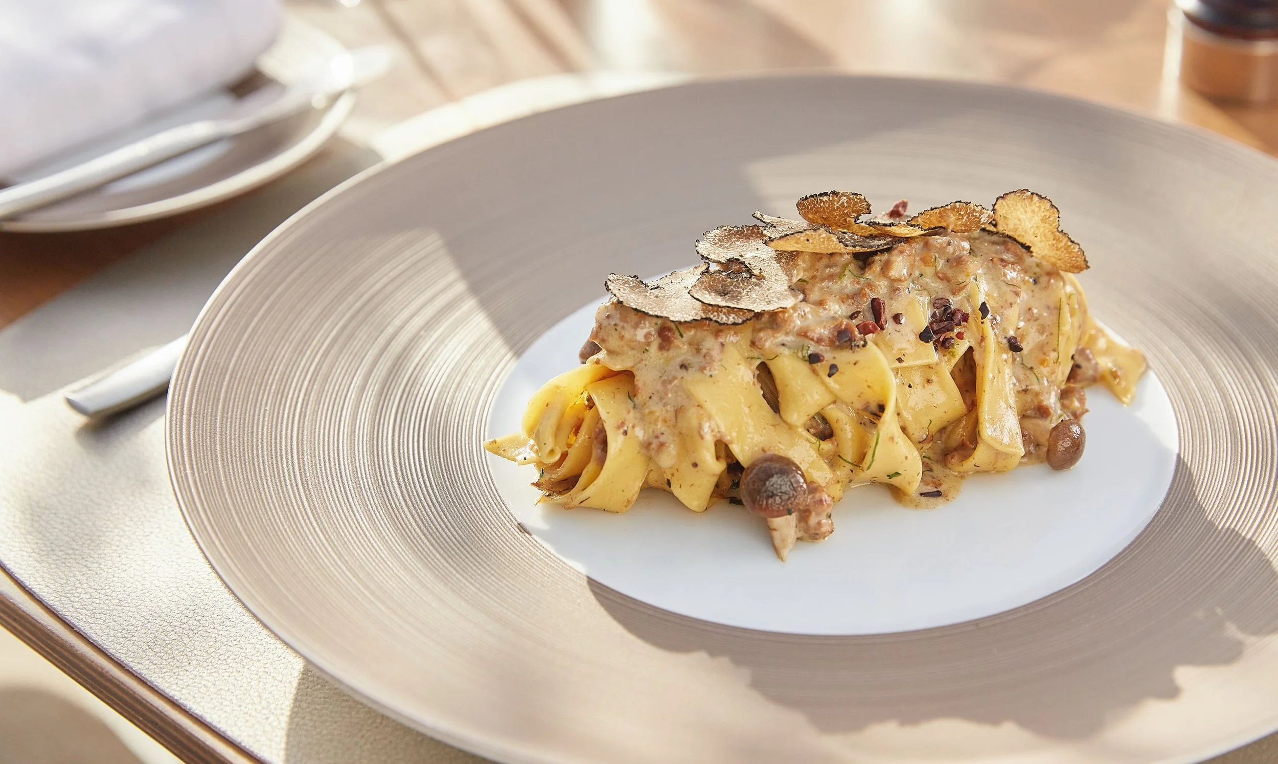 Fettuccine pasta with a creamy sauce, topped with truffle slices and small mushrooms, served on a white plate with a gold-rimmed border.