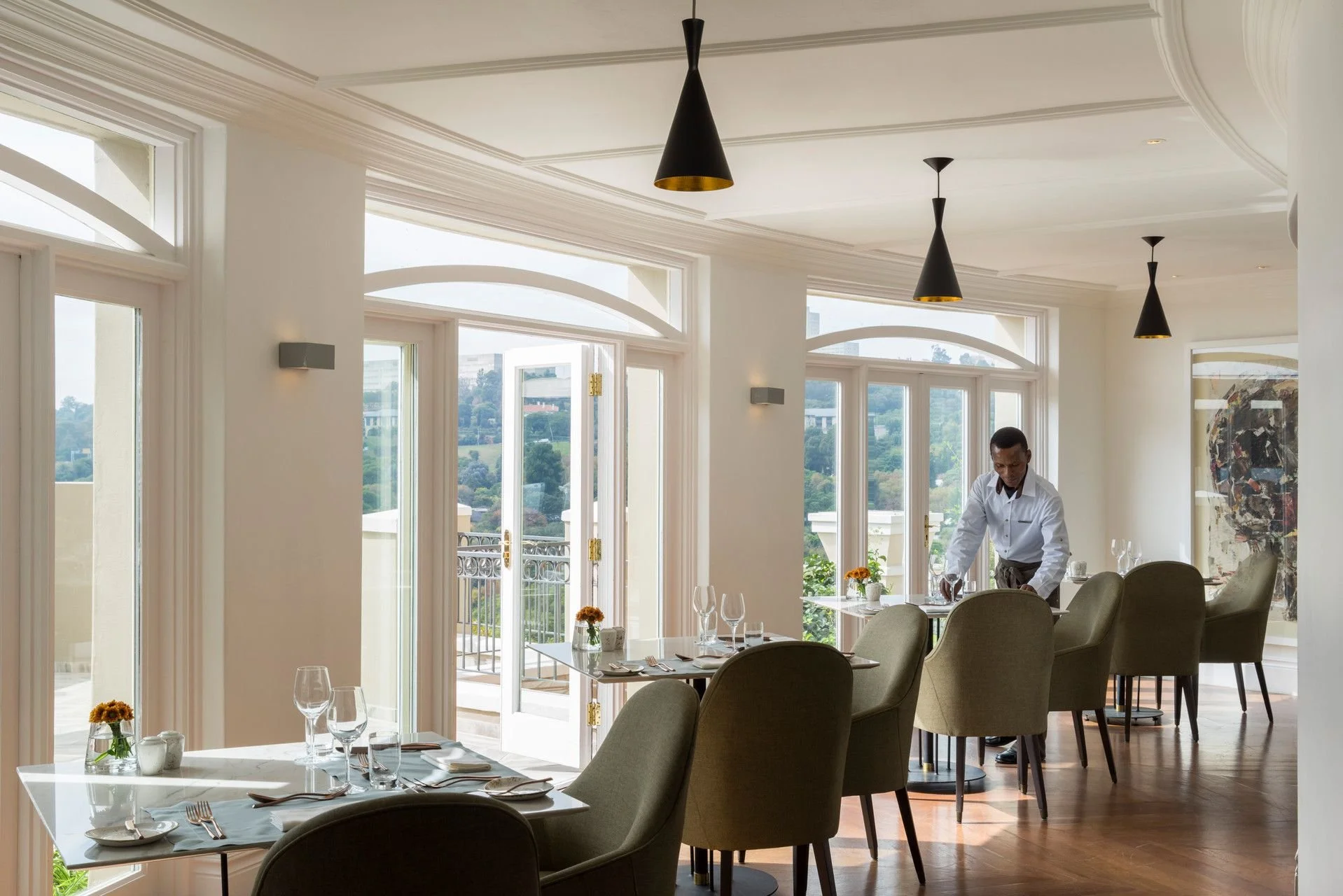 Elegant dining room with large glass windows and a balcony, set with tables, chairs, and wine glasses, featuring a man preparing to serve, and scenery of hills and buildings outside.