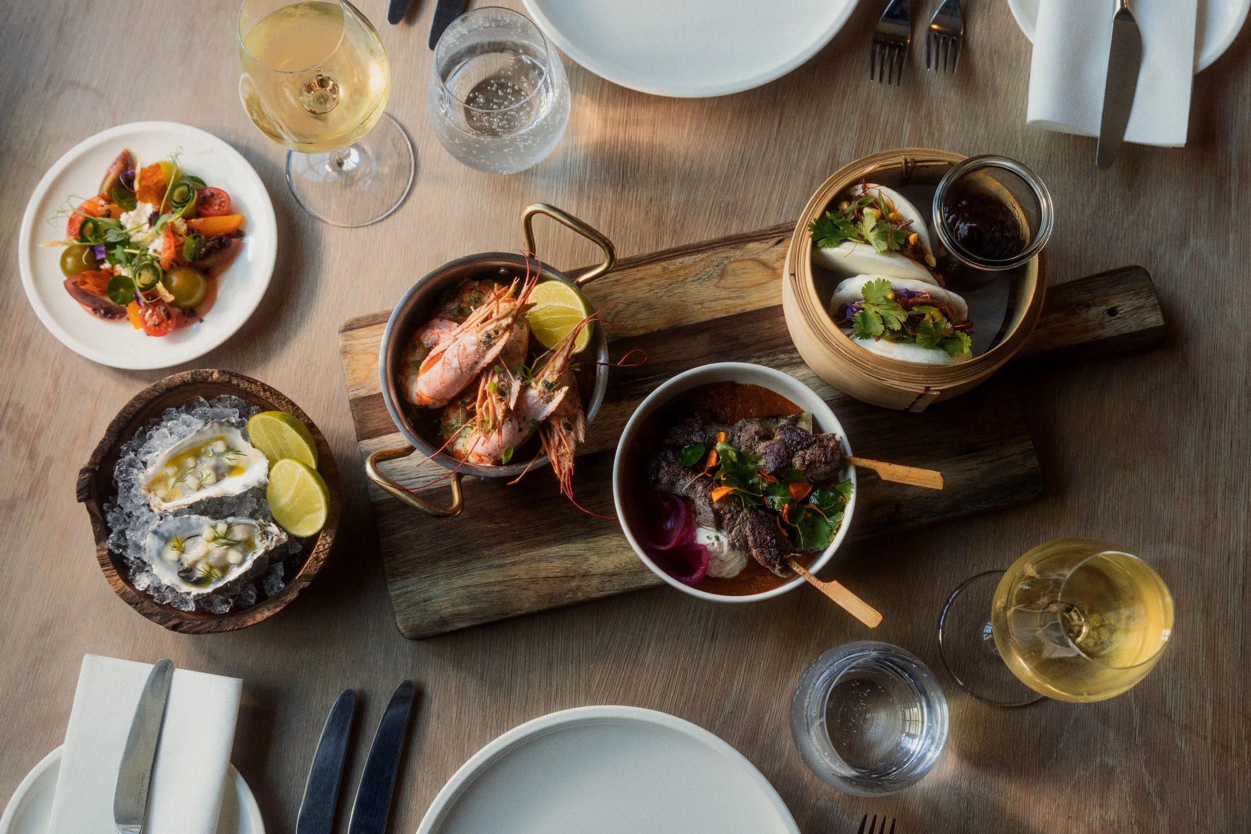 A table with various dishes including a plate of salad, a bowl of oysters with lemon wedges, a seafood dish with prawns, a basket of tacos, and glasses of white wine and water on a wooden table.