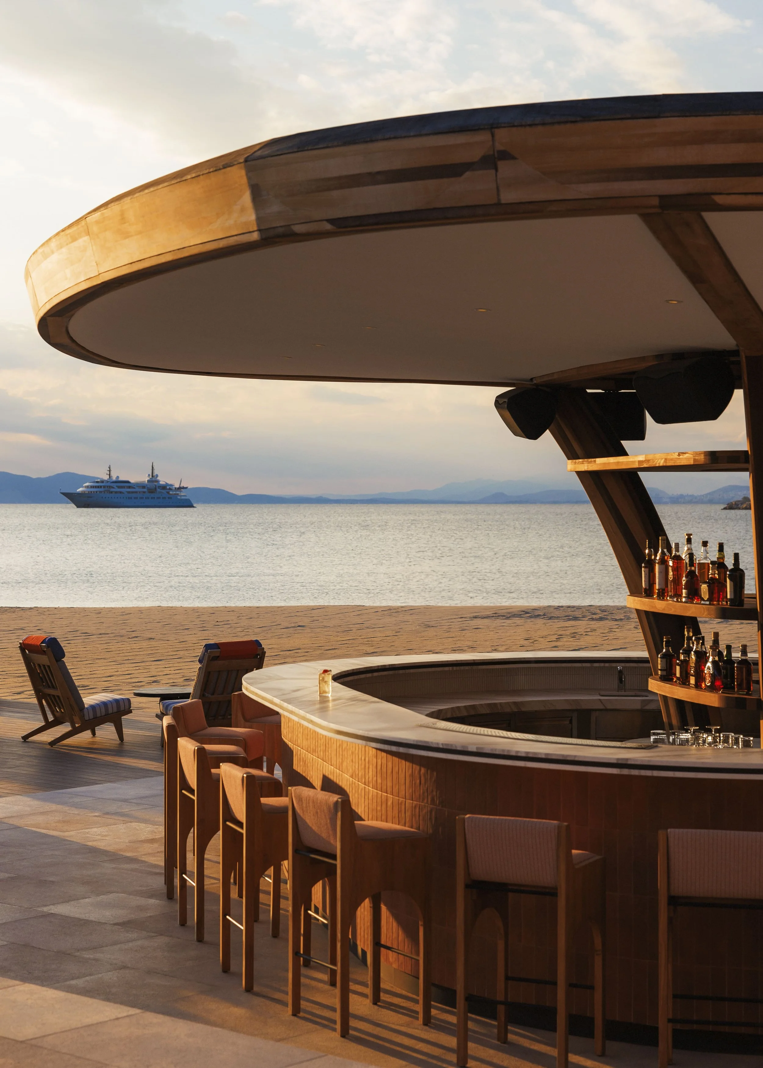 An outdoor bar with a curved white countertop, surrounded by wooden barstools, overlooking a body of water with a cruise ship and mountains in the background during sunset.
