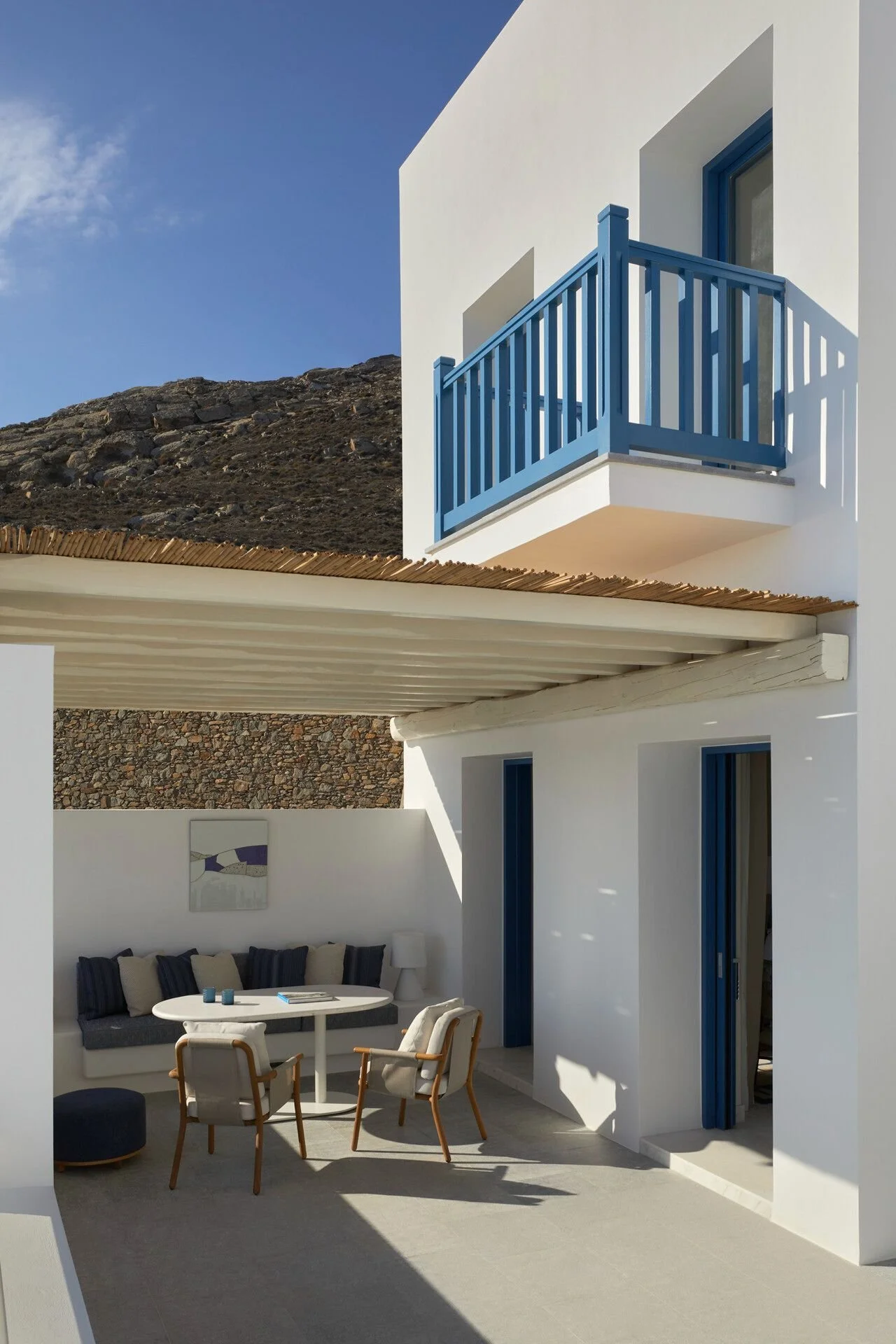A modern white building with blue accents, featuring a small balcony with a blue railing. The outdoor sitting area has a round table with chairs and a cushioned bench with pillows, all under a shaded patio. In the background, a mountain and blue sky are visible.