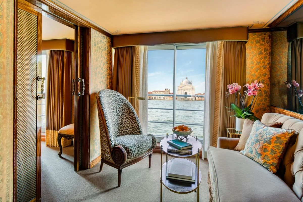 Living room with a view of a cityscape and water, featuring a sofa, an armchair with animal print, a small round table with books and a bowl of fruit, and a potted orchid near the window.
