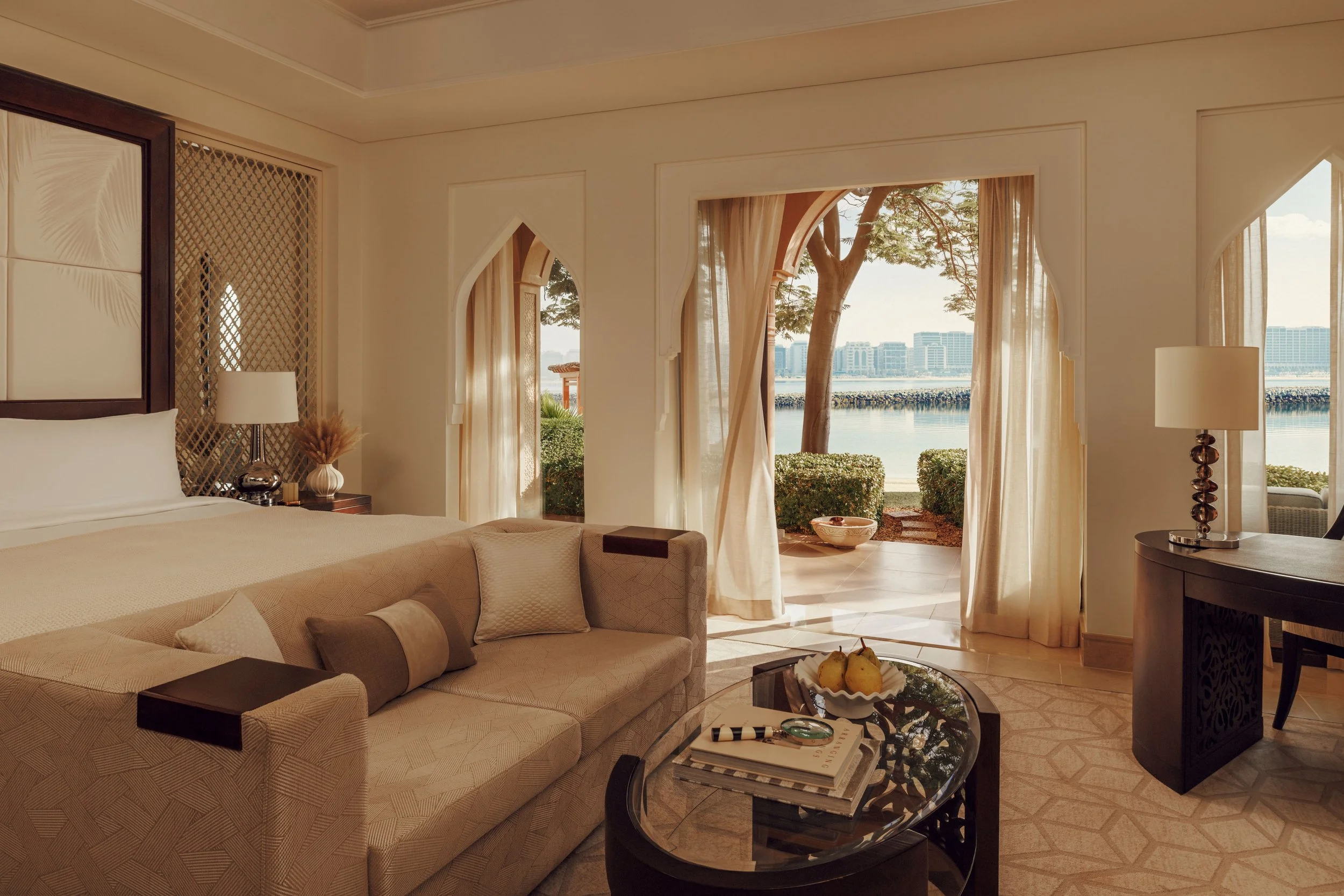 A luxurious hotel suite with a large bed, a cozy sofa, a coffee table with decor, and large windows overlooking a waterfront cityscape with trees.