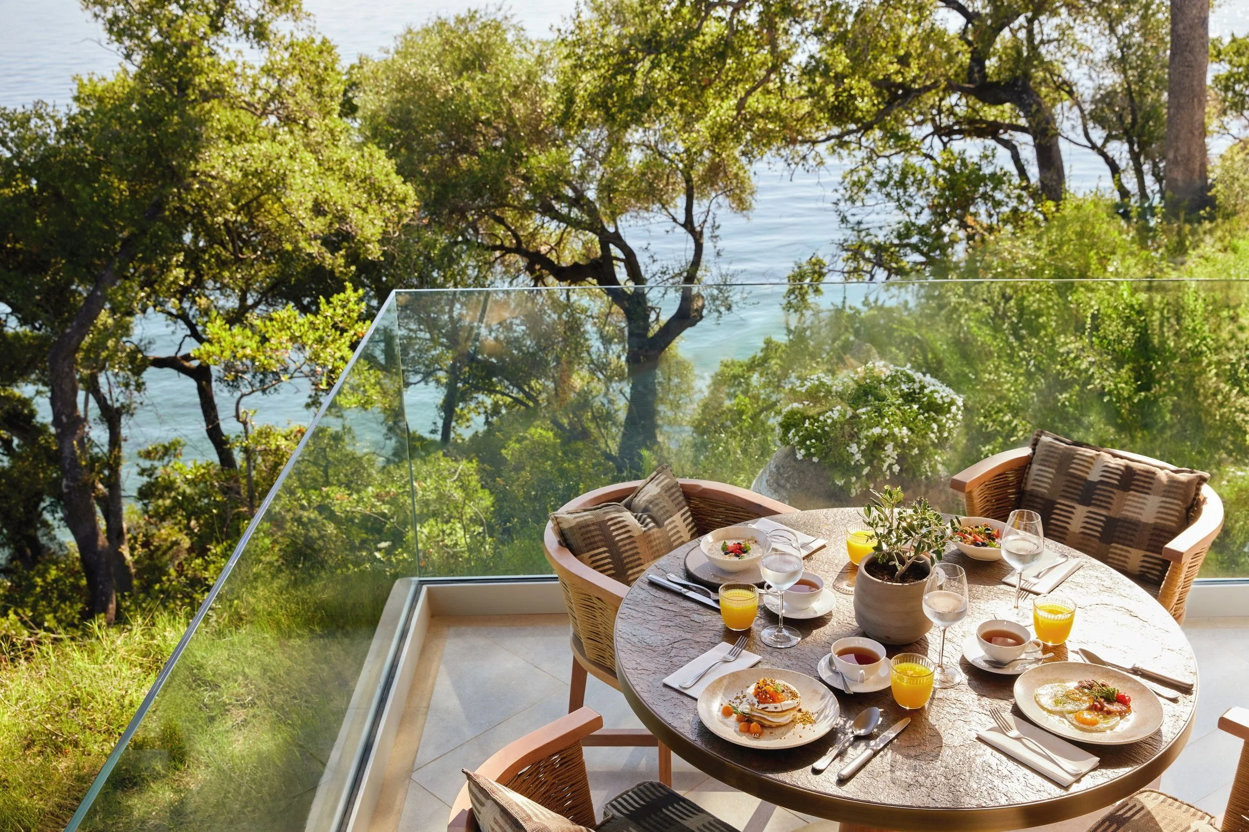 Outdoor dining table set for breakfast with bowls of food, glasses of orange juice, and silverware, on a patio overlooking lush green trees and water.