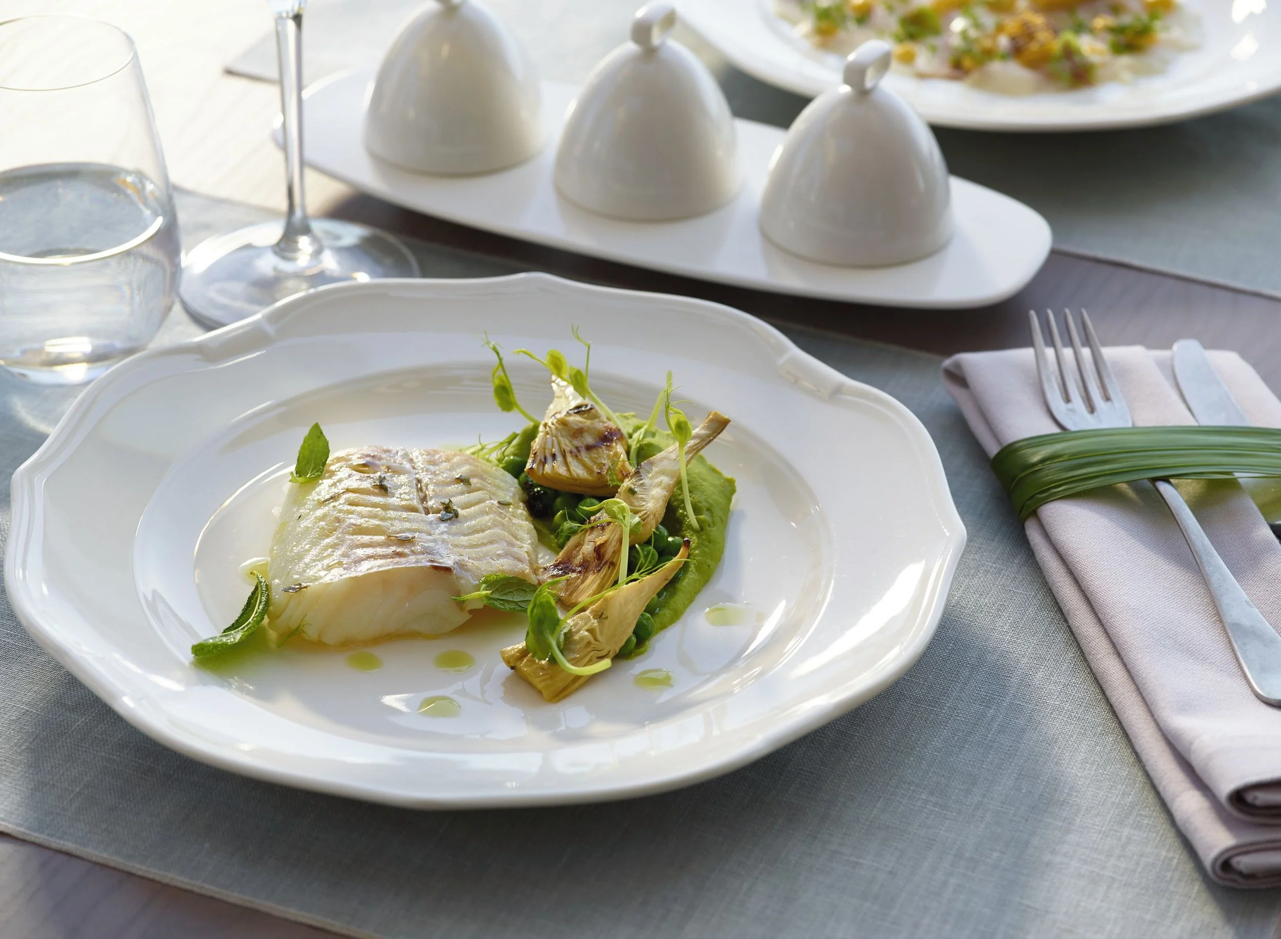 A white plate with a cooked fish fillet topped with herbs and served with grilled artichoke hearts and microgreens. A glass of water, a wine glass, a set of three small white condiment cups, and a fork with a napkin wrapped with a green ring are also on the table. There is a grassy salad on a separate plate in the background.