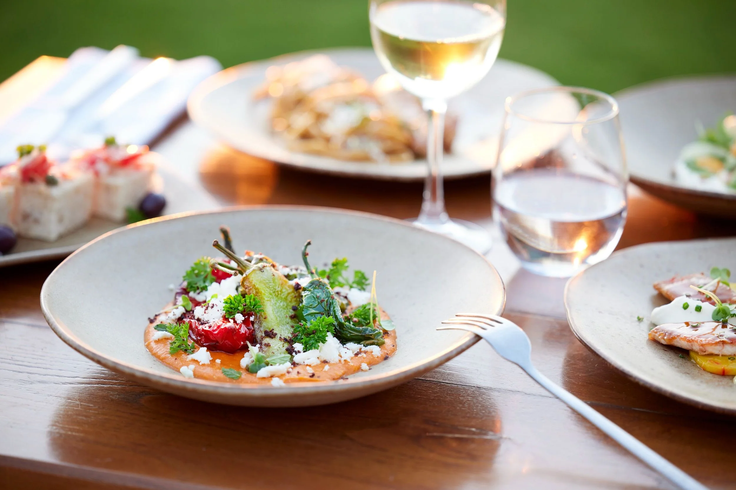 Close-up of a plate with an elegantly garnished appetizer featuring greens, cheese, and roasted vegetables on a wooden table with wine glasses and other dishes in the background.