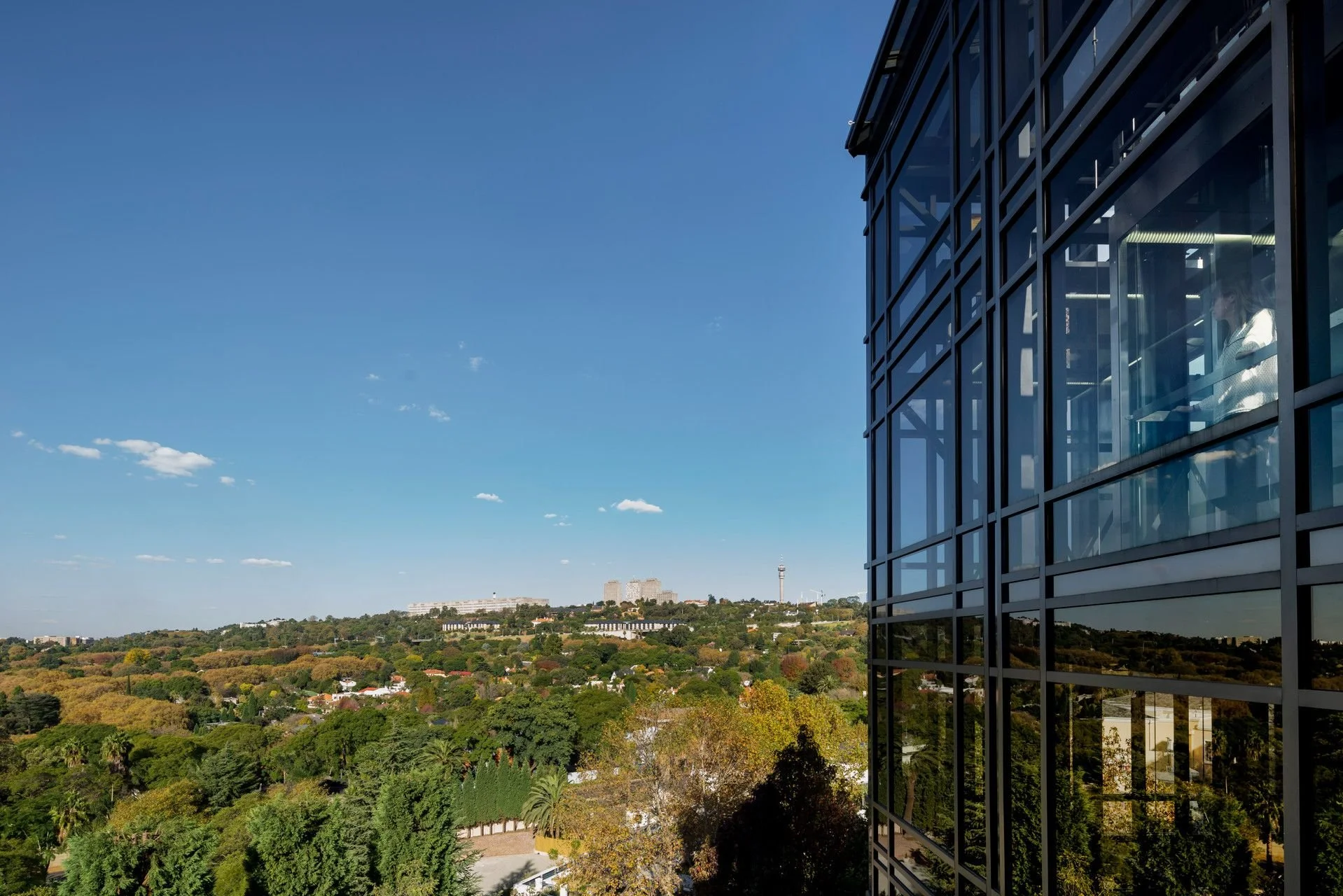 View of a glass building on the right with a scenic landscape of trees, houses, and distant city buildings under a clear blue sky.