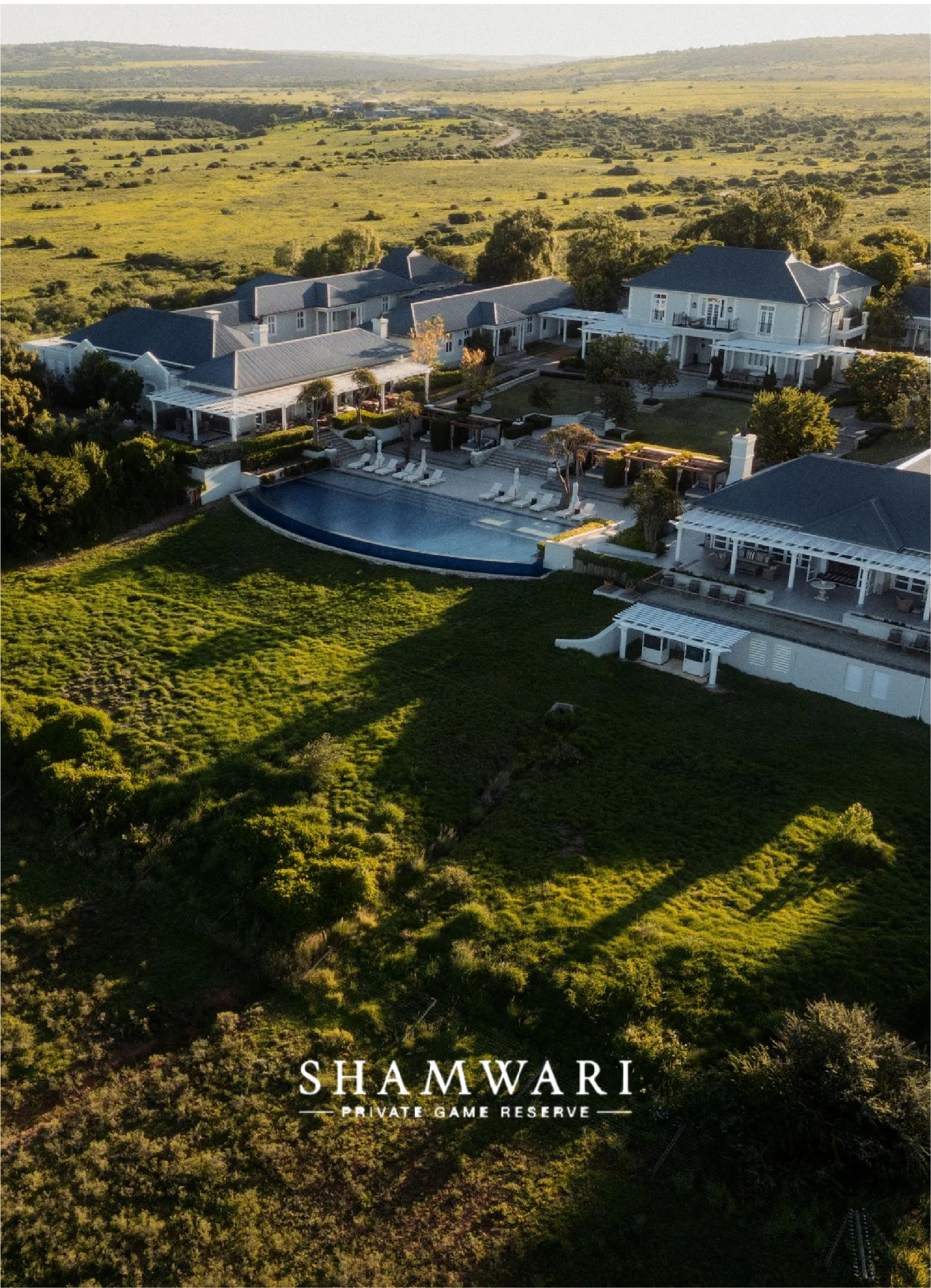 Aerial view of a luxurious estate called Shamwari Private Game Reserve with multiple large buildings and a swimming pool, surrounded by lush green vegetation and open plains.