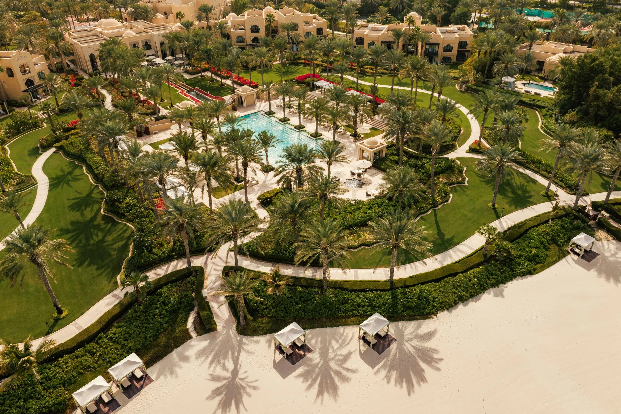 An aerial view of a luxurious hotel resort featuring a large swimming pool, surrounded by palm trees, green lawns, pathways, and small cabanas on a sandy beach.