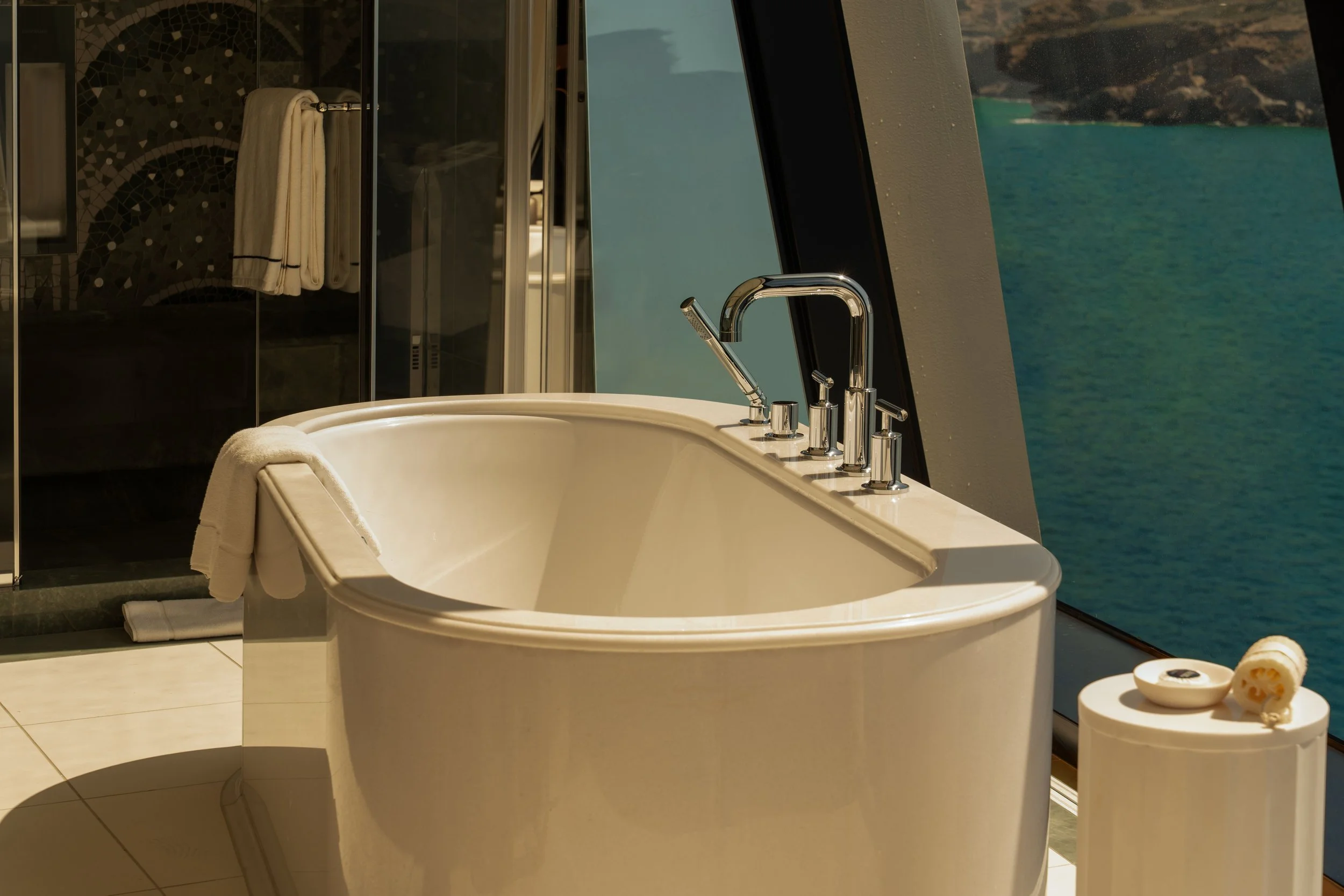 A luxurious bathtub with a view of the ocean through large floor-to-ceiling windows. The bathtub has silver fixtures and a soft towel on its edge. Next to the tub, there are spa items including bath bombs, soap, and a rolled-up towel. The room features dark tiles, a glass-enclosed shower, and a scenic coastal landscape visible outside.