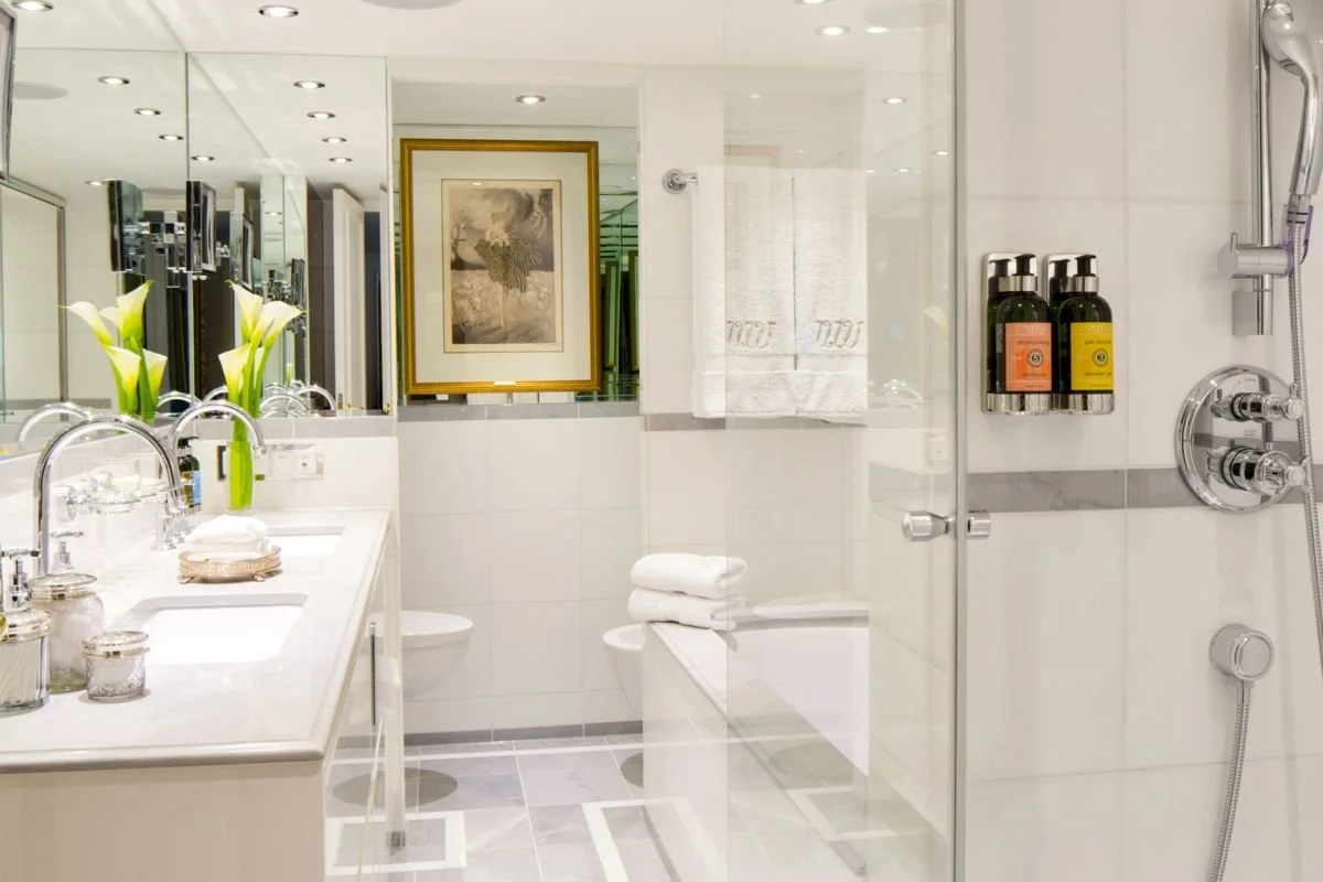 Modern bathroom with double sink vanity, white towels, framed artwork, and a shower with bottles of soap and shampoo