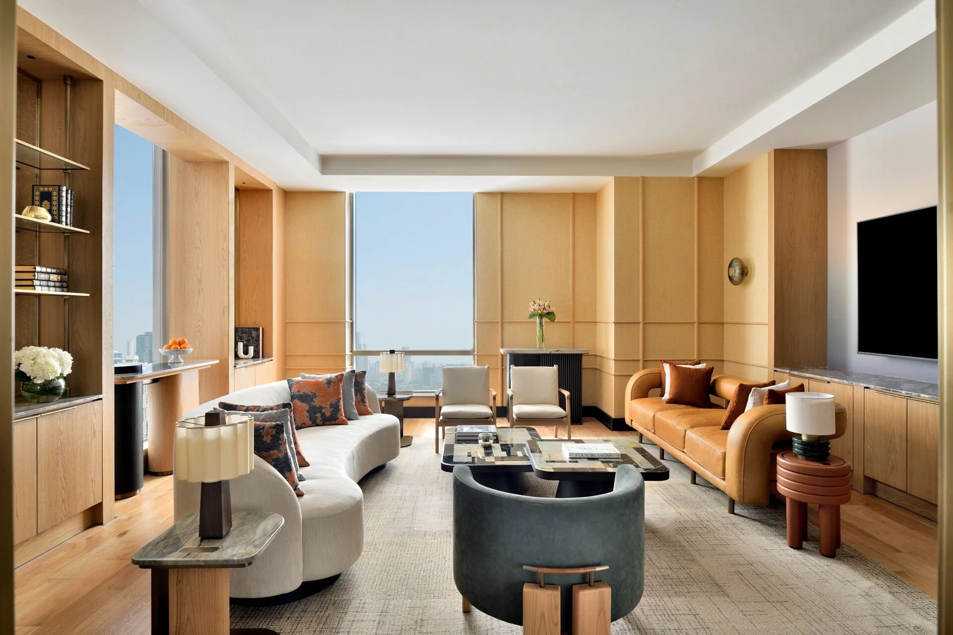 Modern living room with beige walls, large windows, and mid-century furniture including a white sofa with patterned pillows, a tan leather sofa, and several armchairs. Decor includes a coffee table, lamps, books, and flowers.