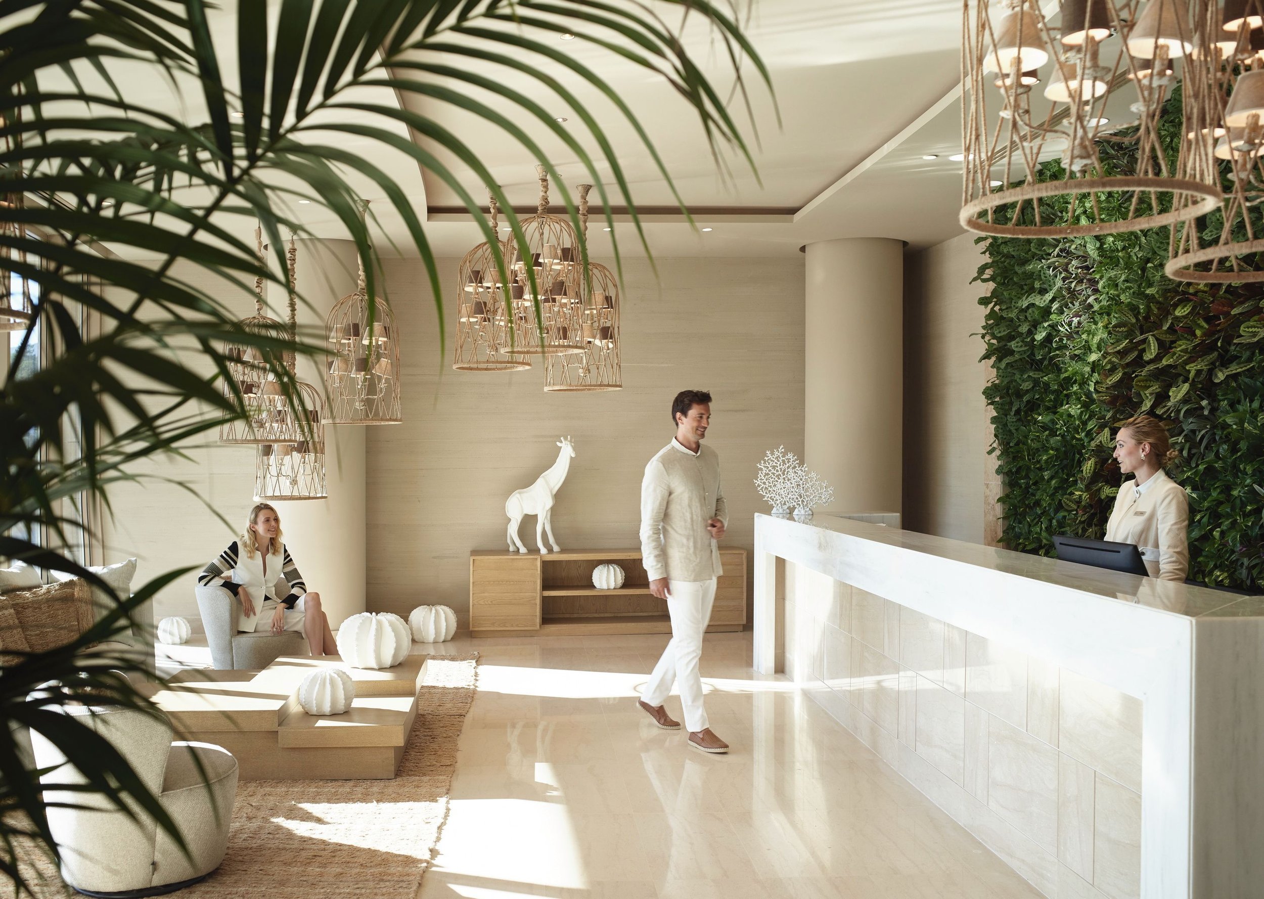 A modern hotel lobby with natural light, greenery, and decorative lighting fixtures. There are three people, two women seated and a man standing near the reception desk, with white decor elements such as a horse sculpture and round ornaments.