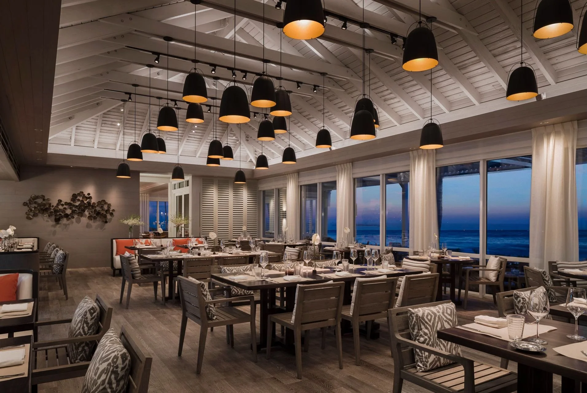 Elegant restaurant interior with wooden tables and patterned chairs, large windows showing a view of the ocean at dusk, and black pendant lights hanging from a white, vaulted ceiling.