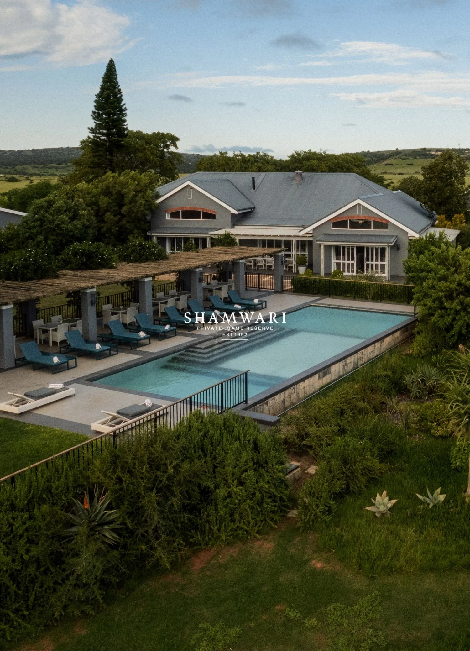 A large house with a gray roof, surrounded by trees and greenery, overlooking rolling hills under a partly cloudy sky. A swimming pool with lounge chairs and an outdoor dining area are in the foreground, and the words 'SHAMWARI PRIVATE GAME RESERVE EST 1992' are overlaid on the image.