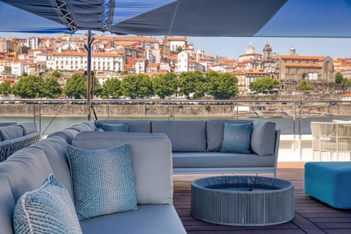 Outdoor patio with white sofas and blue cushions, large blue umbrella, overlooking a cityscape with historic buildings and a river.