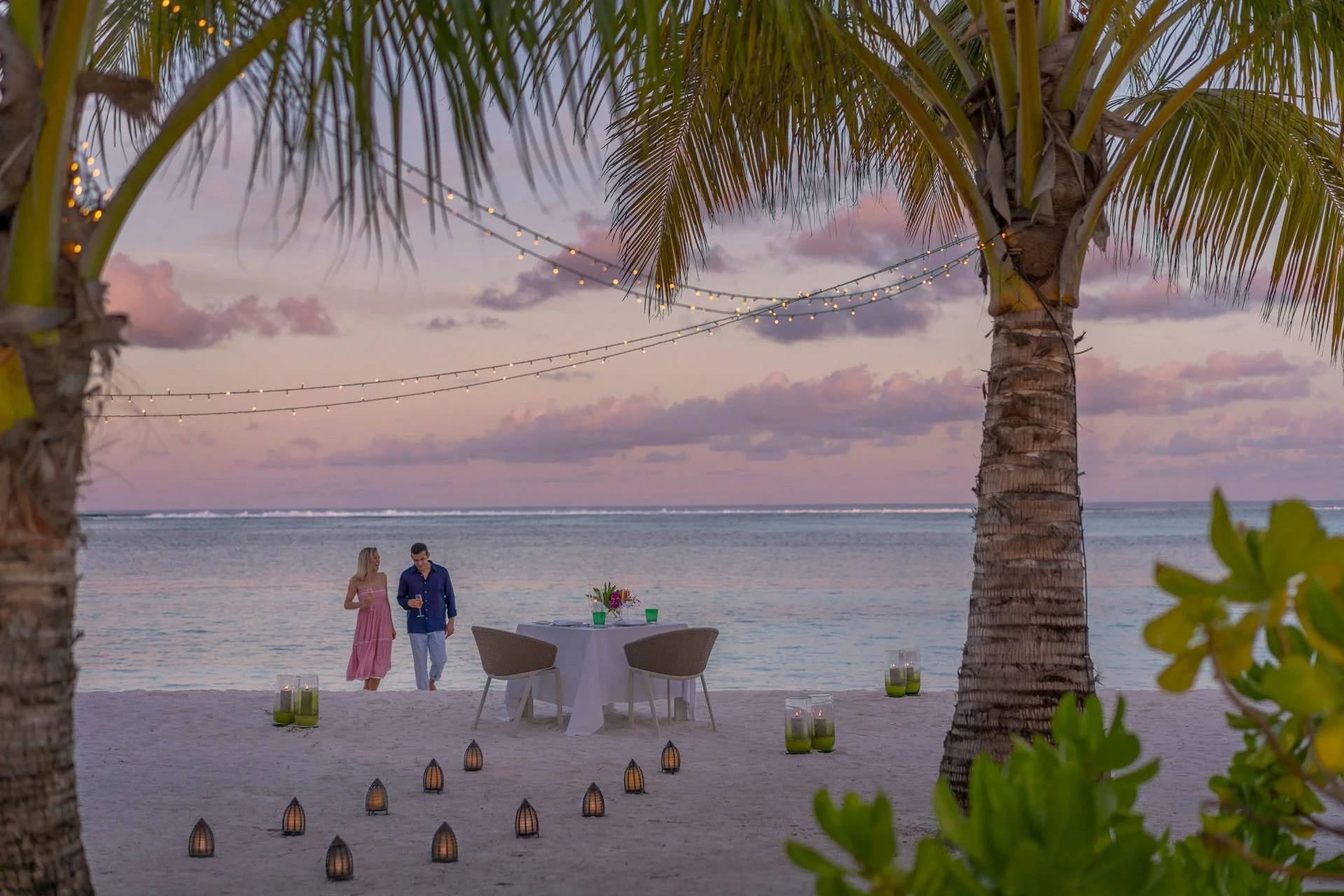 A beach setting at dusk with two people standing near a table decorated with flowers, surrounded by palm trees and lanterns in the sand, with a pink and purple sky reflected on the calm ocean.
