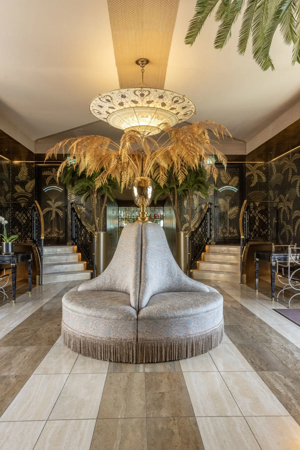 Elegant hotel lobby with a circular beige upholstered seating area, large gold vase with dried palm leaves, and stairs with black railings leading to an upper level, decorated with gold palm-frond wall art and a decorative ceiling fixture.