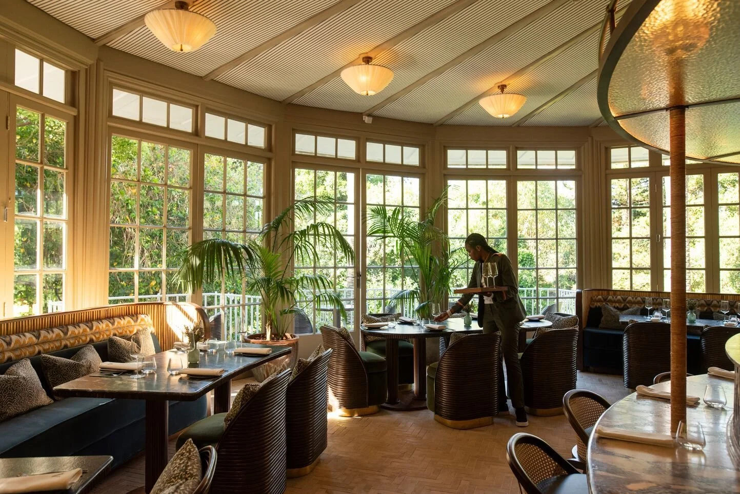 Upscale restaurant interior with large windows, potted plants, and a server setting a table.