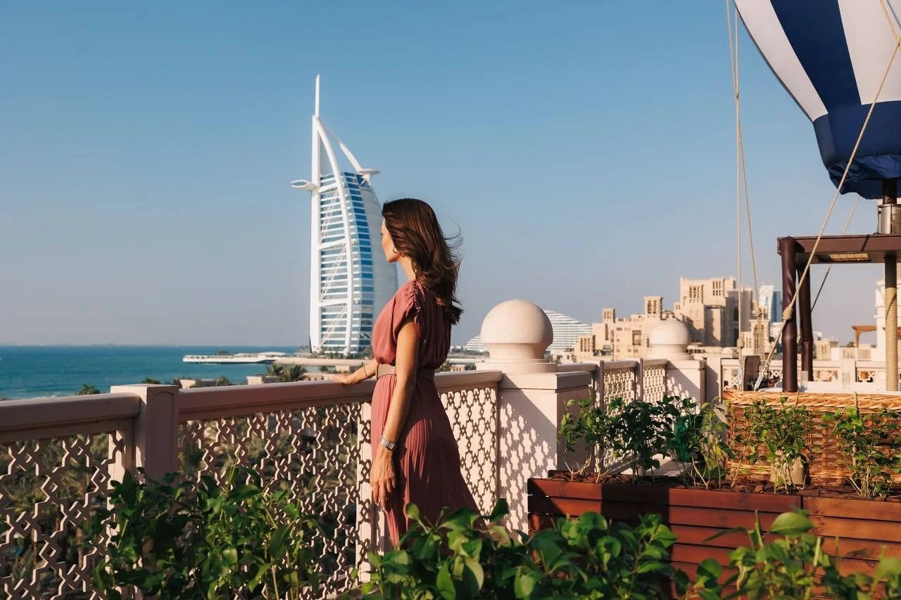 Woman standing on a balcony overlooking the cityscape of Dubai, including the Burj Al Arab hotel and the Arabian Gulf.