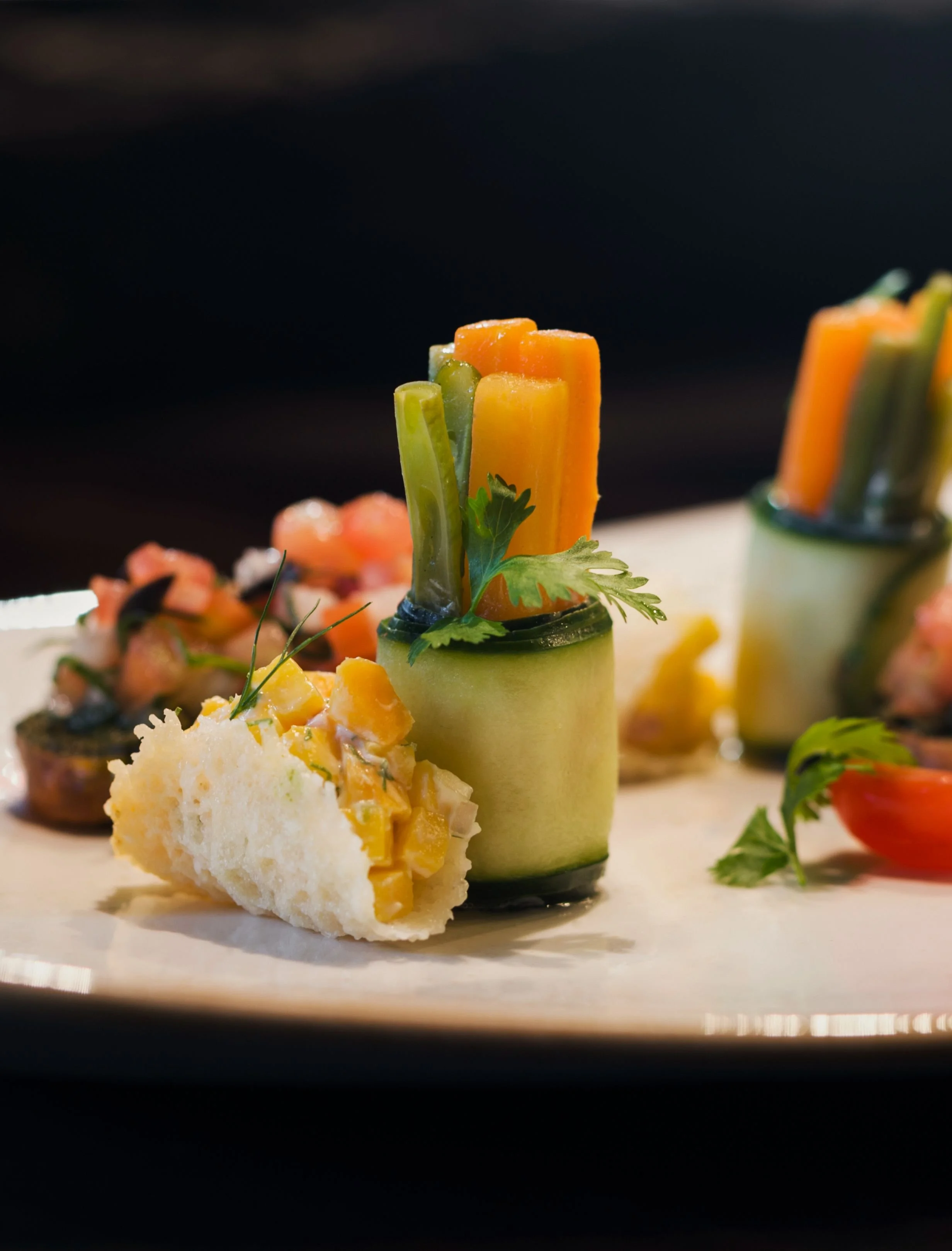Close-up of vegetable appetizers, including cucumber roll with carrot and celery sticks, garnished with a sprig of parsley, served on a white platter.