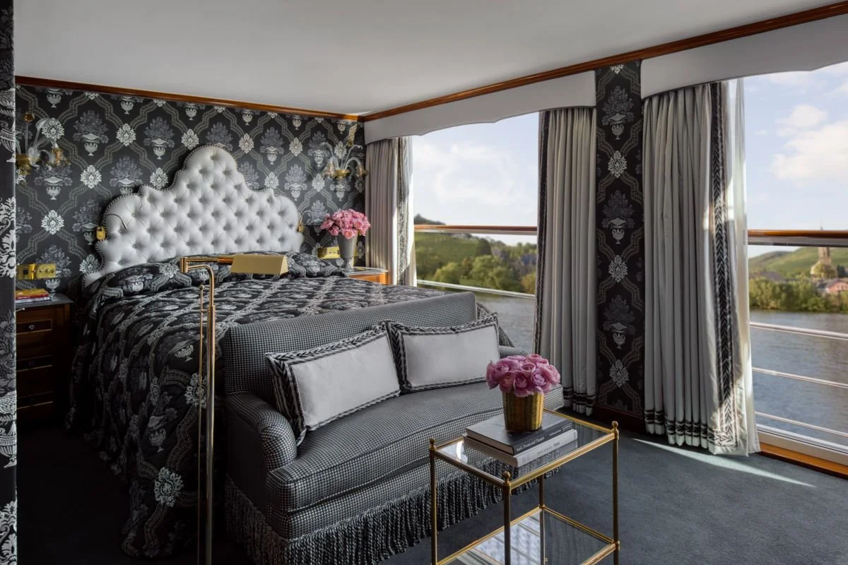 Luxurious bedroom with a large tufted white headboard, black and white damask patterned wallpaper, and a view of a river and green landscape outside large windows with cream curtains.