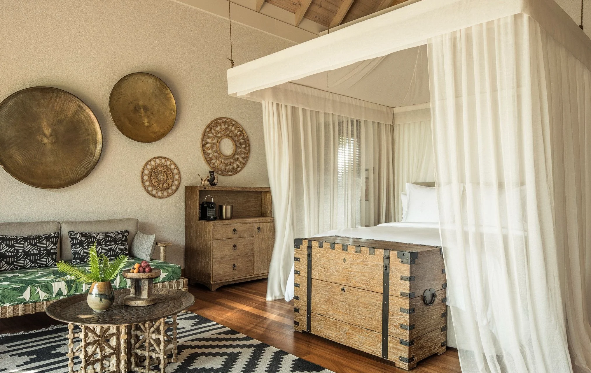Living room with a beige sofa, decorative pillows, a plant on a round wicker table, a wooden cabinet with a coffee machine and cups, wall decor including circular metal art, and a four-poster bed with curtains and a wooden trunk at the foot of the bed.