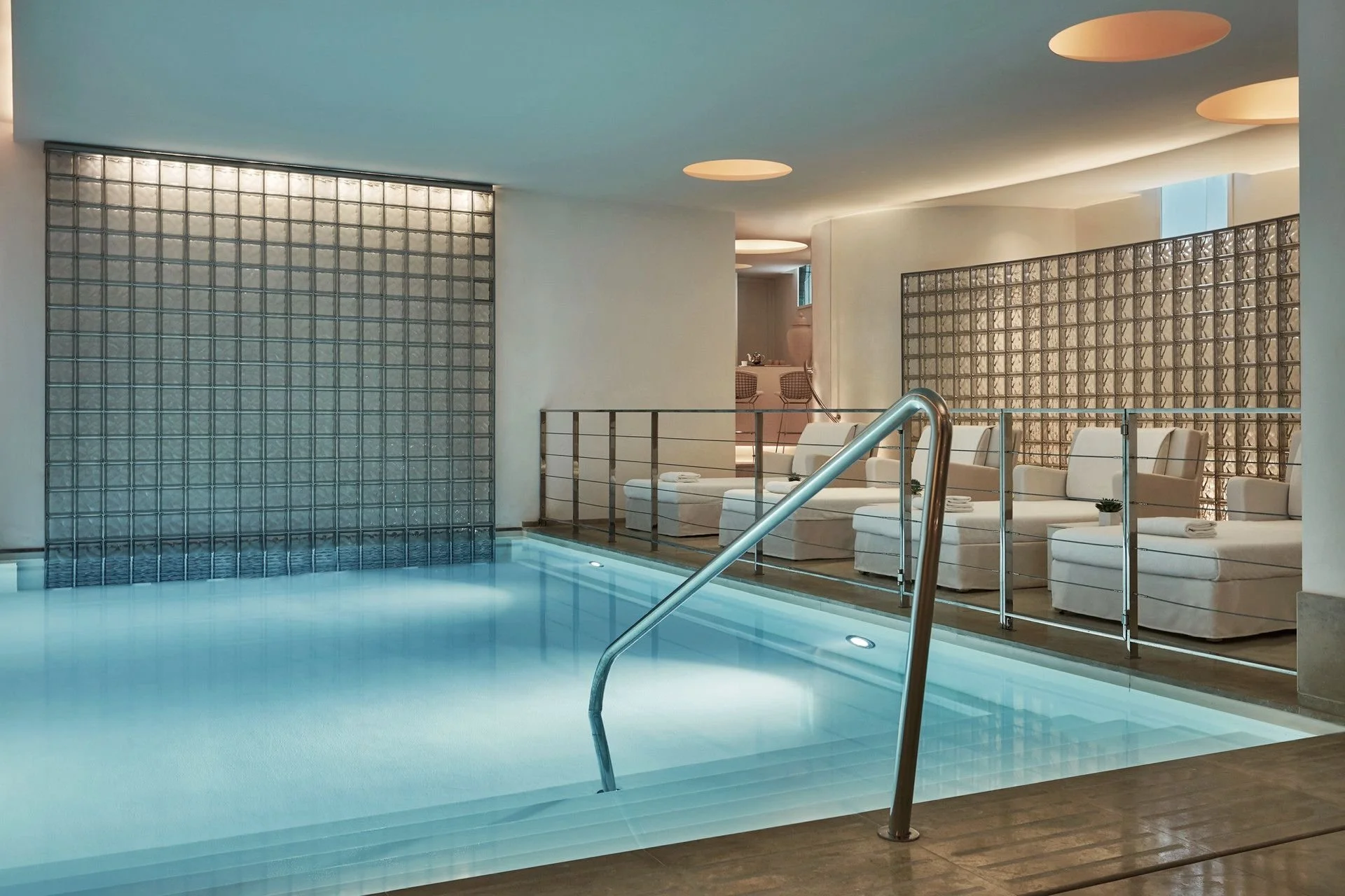 Indoor swimming pool area with a row of white lounge chairs and a decorative glass brick wall in the background.