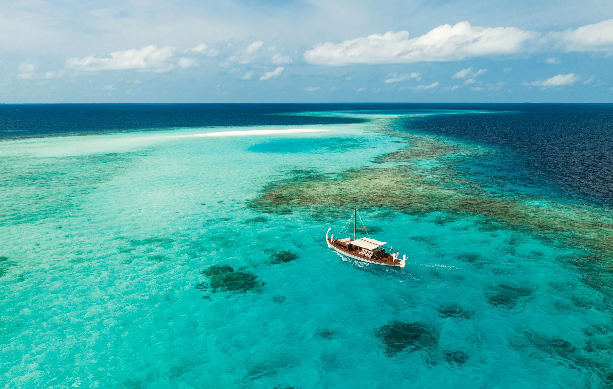 A sailboat floating on clear turquoise water near a coral reef, with a partly cloudy sky overhead.
