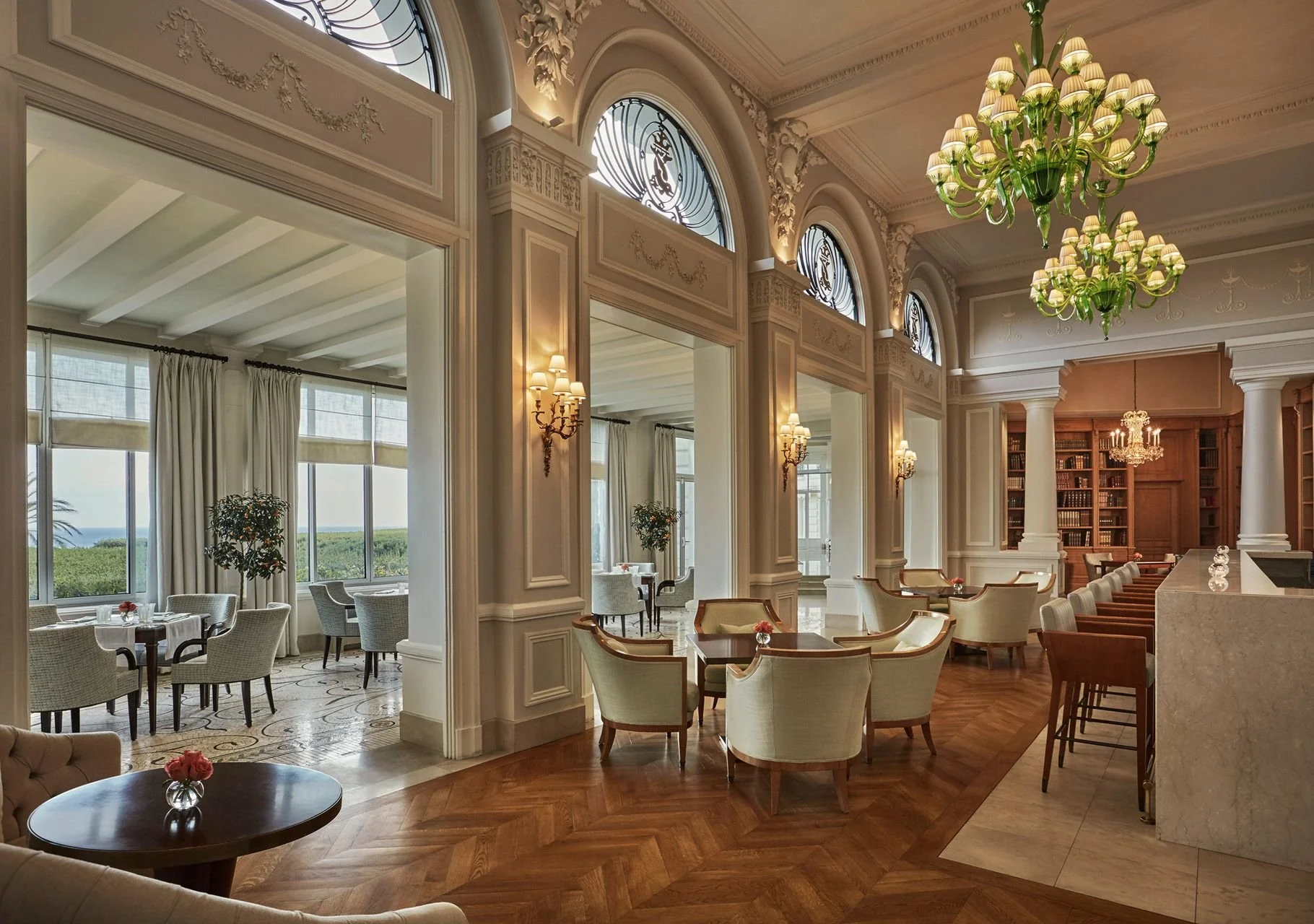 Elegant hotel or restaurant interior with high ceilings, ornate moldings, large arched windows, and multiple chandeliers. The room features a bar area with bar stools, a library with tall bookshelves, and seating arrangements including armchairs and small tables, creating a luxurious and comfortable atmosphere.