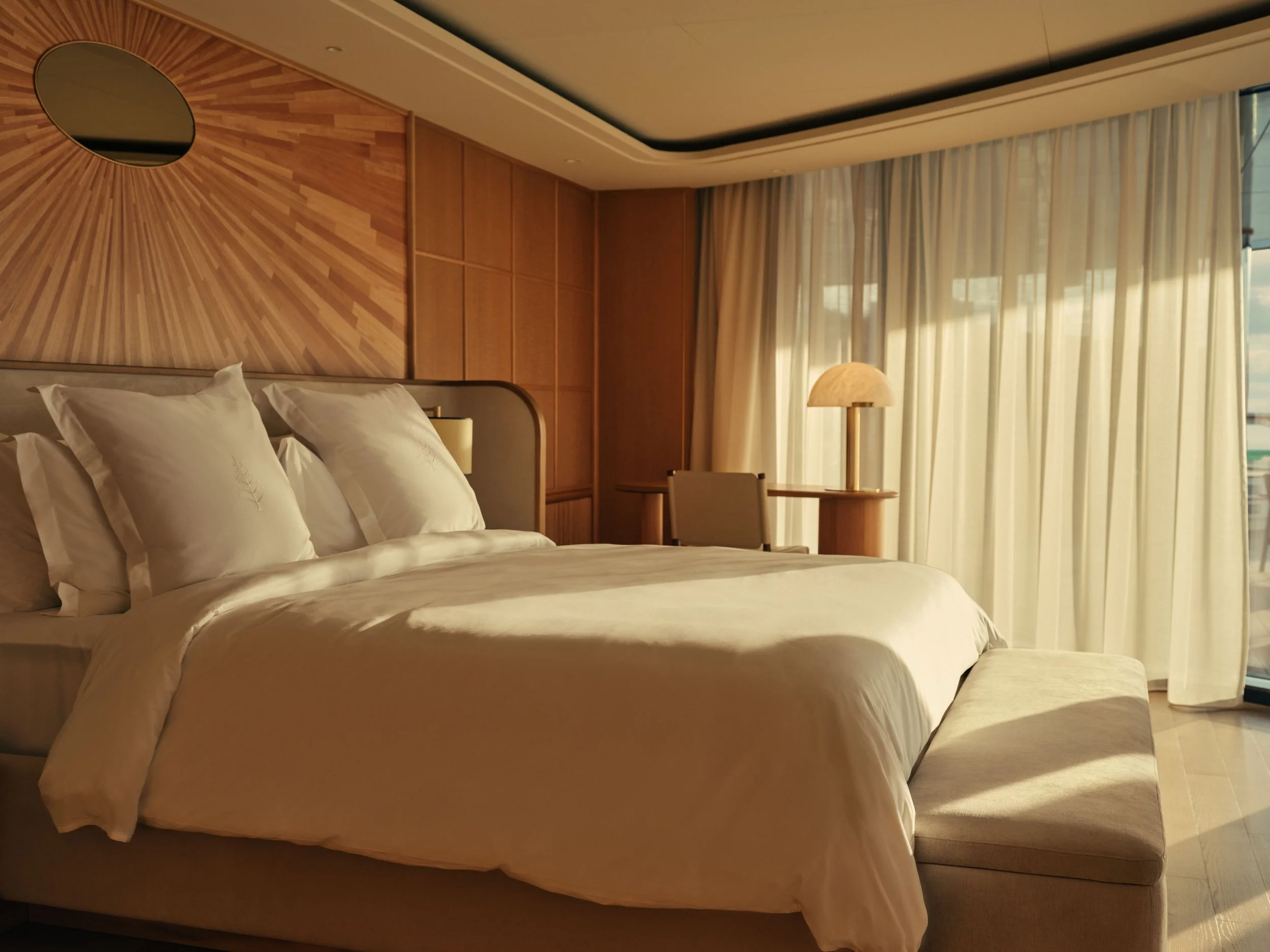 A hotel room with a large bed, white pillows, a wooden headboard, a bedside lamp, and a seating area by large windows with sheer curtains.
