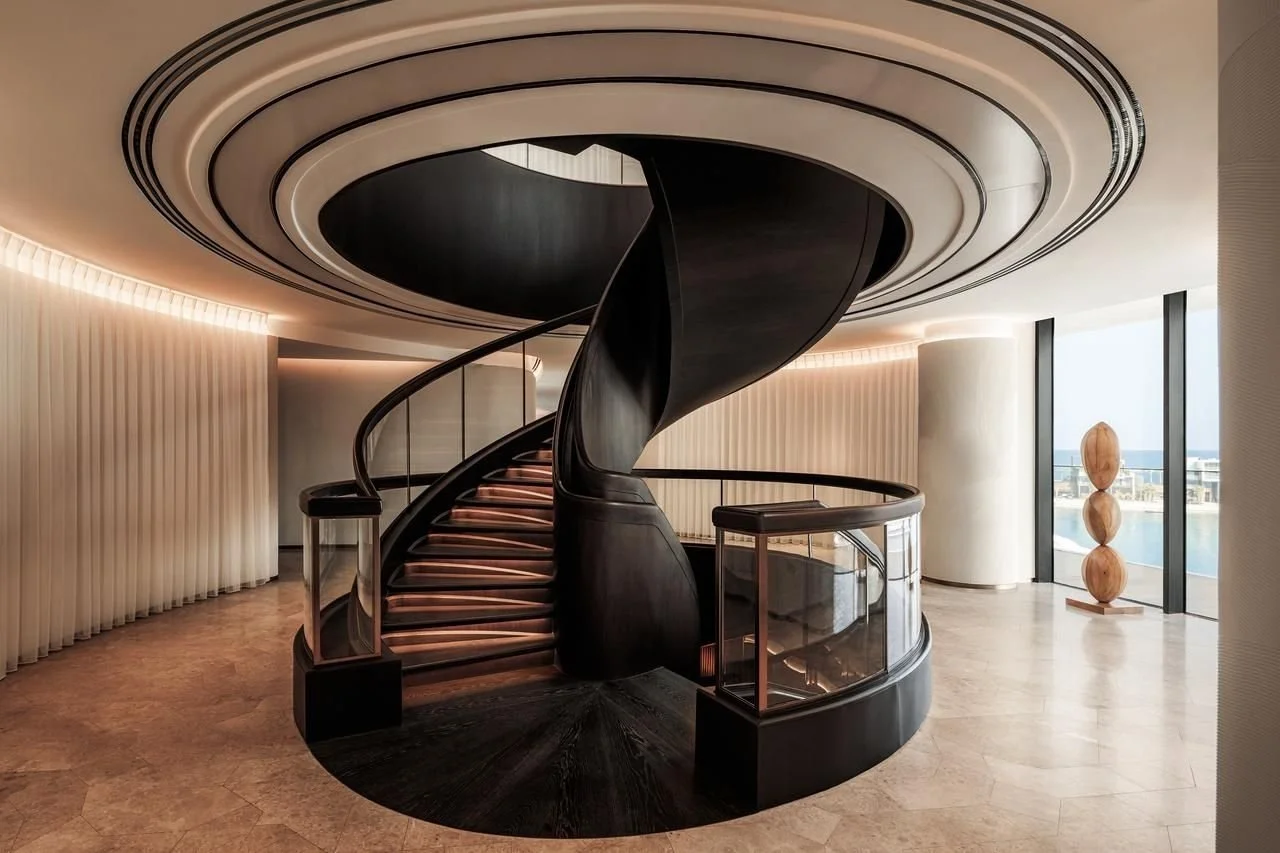 Elegant interior of a modern building with a black spiral staircase, large windows with a city view, and a sculpture of three stacked wooden spheres.