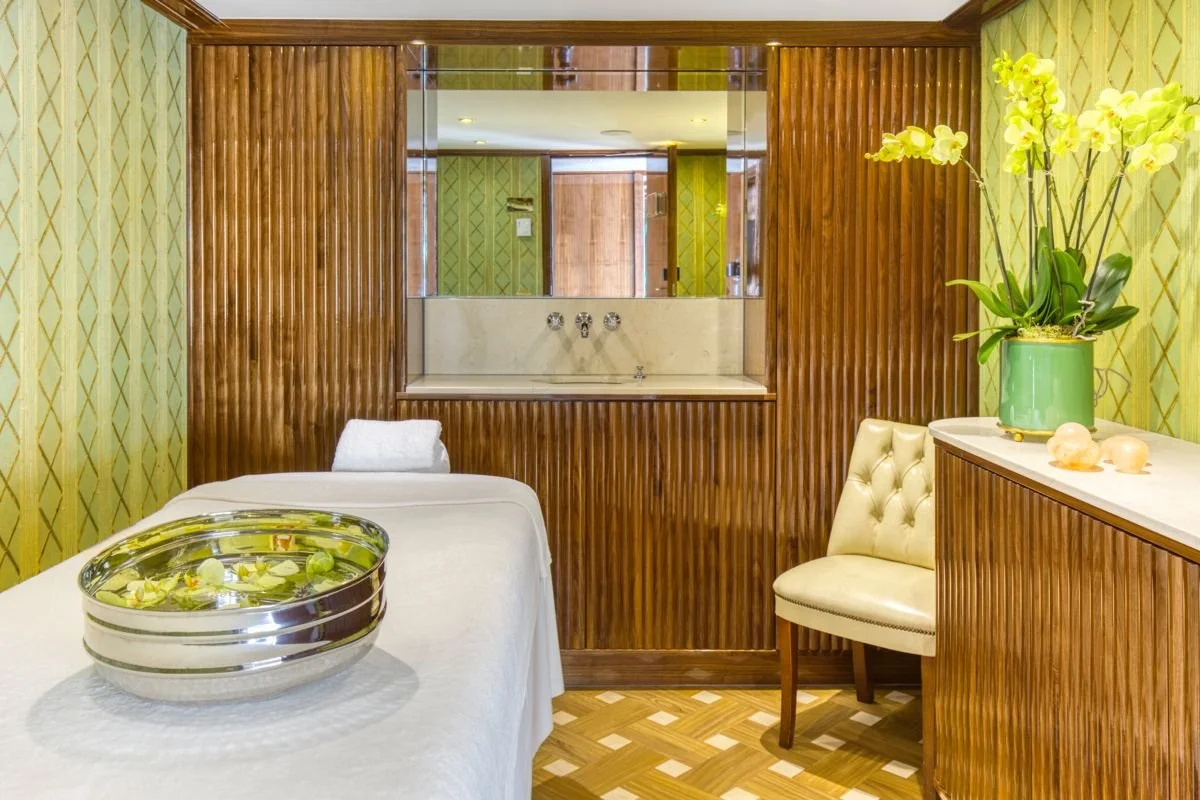 A spa or massage room with a massage table covered in white linens and a towel. There is a metal bowl with water and floating flowers on the bed. The room has wooden paneling, a mirror, and a marble sink. A white upholstered chair and a vase with yellow orchids are also present.