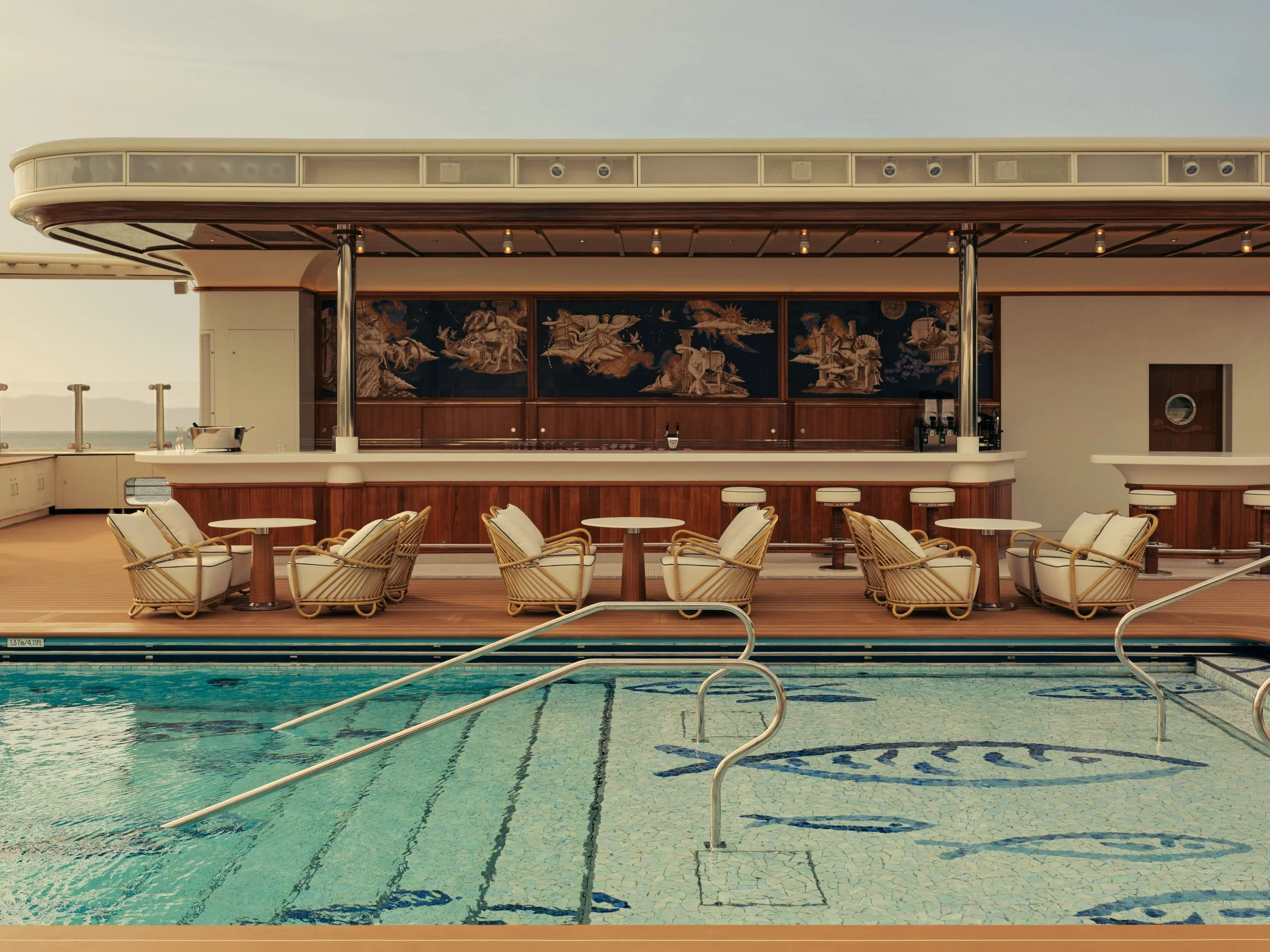 Empty poolside lounge chairs and tables with a swimming pool in the foreground, and a bar area with artwork and ceiling lights in the background.