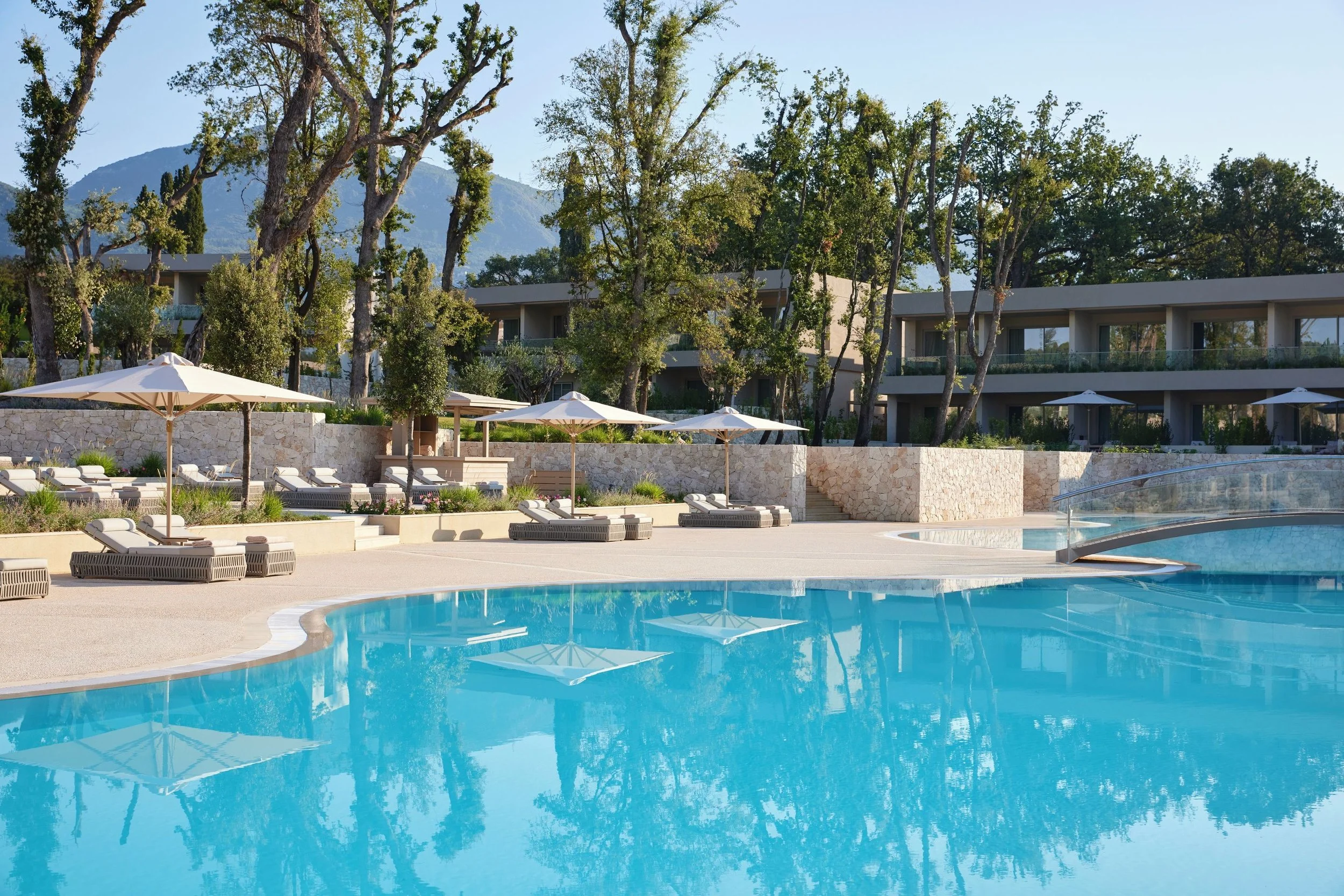 A swimming pool area with lounge chairs and umbrellas, surrounded by trees and modern buildings, with mountains in the background.
