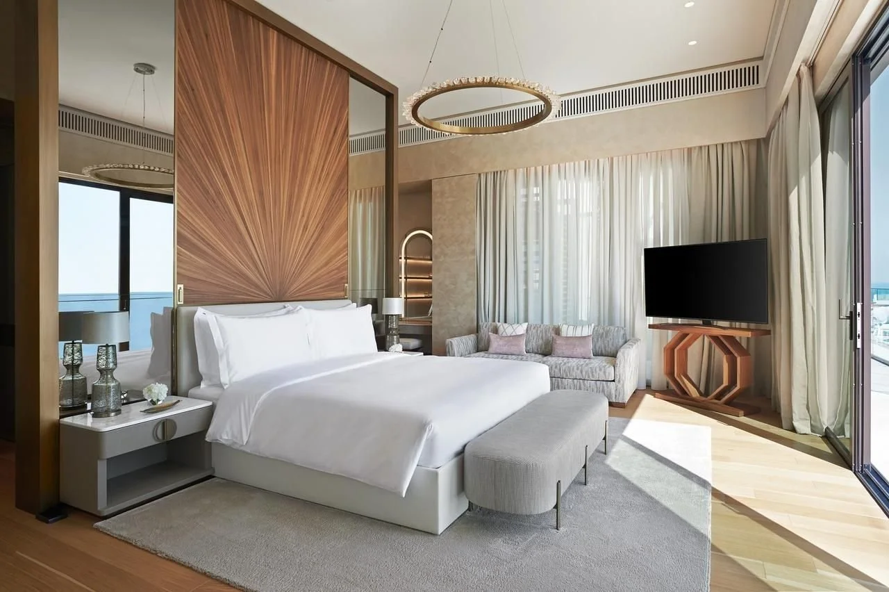 A modern hotel room with a king-size bed, bedside tables with lamps, a gray upholstered footstool, a large flat-screen TV on a wooden stand, a sitting area with a sofa and pink pillows, and large windows with sheer curtains allowing natural light.