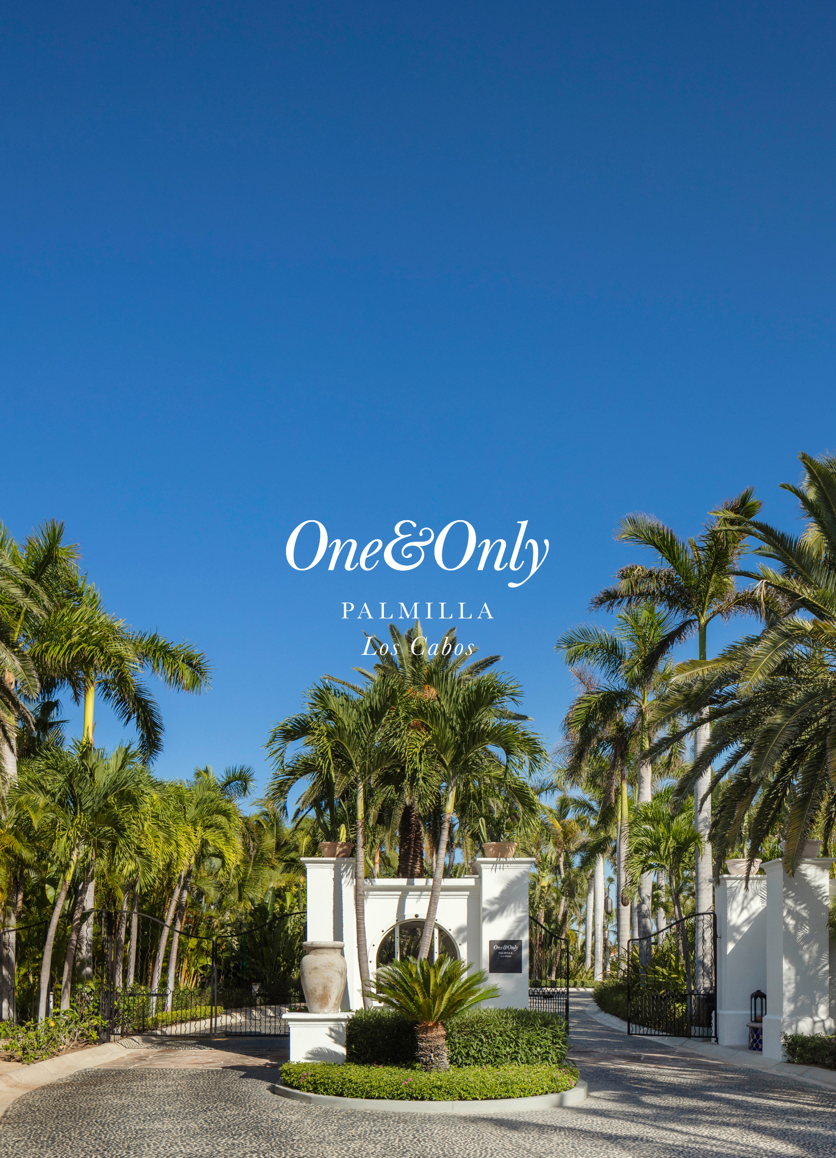 Entrance to One&Only Palmilla resort in Los Cabos, with white walls, a white archway, palm trees, and a clear blue sky.
