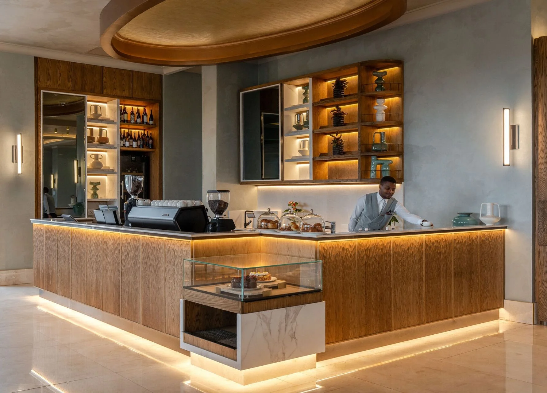 A modern hotel or cafe counter with wooden paneling and warm lighting, a barista preparing drinks, and decorative shelves with vases and bottles behind the counter.