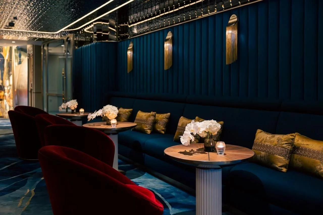 Elegant lounge area with plush red chairs, a long blue velvet sofa with gold and green pillows, round wooden tables with white flowers and votive candles, blue textured wall with gold accents, and a patterned carpet.
