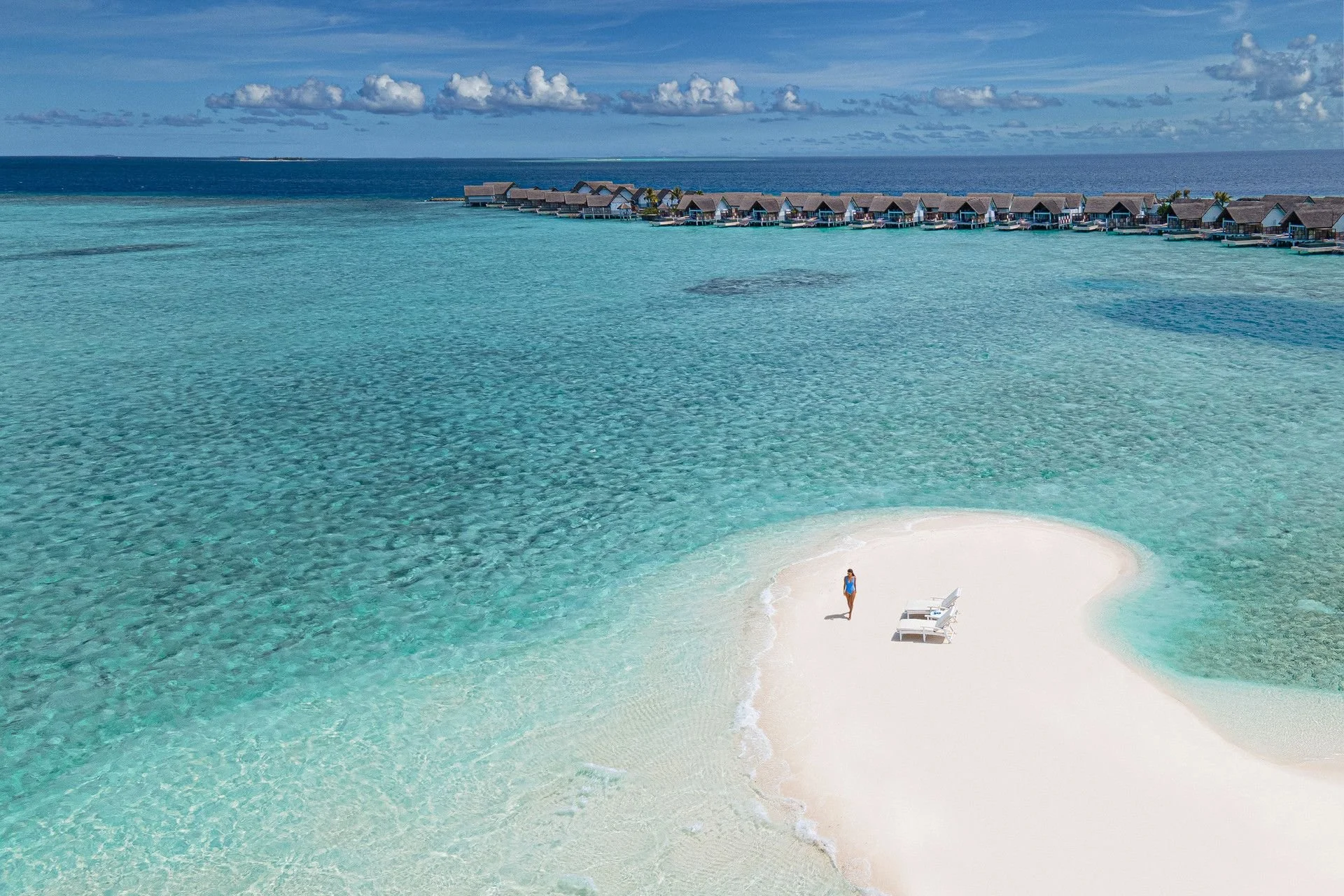 A person standing on a small white sand island with two lounge chairs, surrounded by clear turquoise water, with overwater bavilions in the background and a cloudy sky.