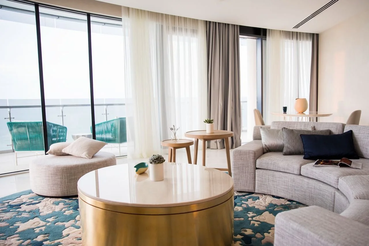 Modern living room with large windows, light-colored sofa with pillows, a round coffee table, small side tables with plant decorations, and a balcony with chairs.