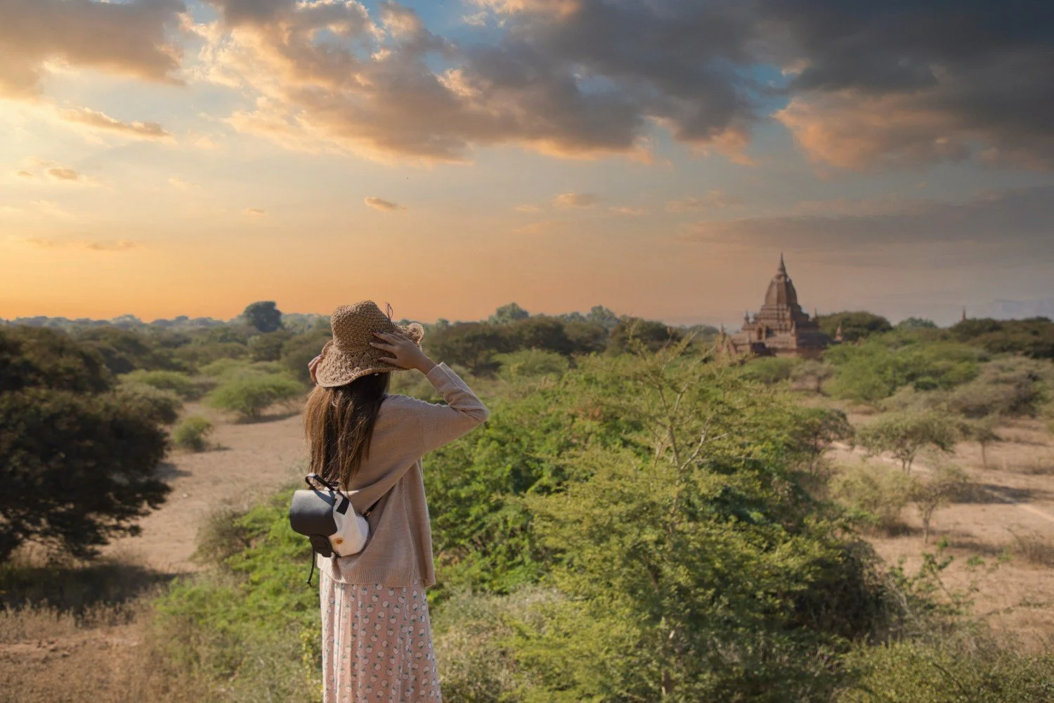 A woman wearing a sunhat and a polka dot skirt stands amidst greenery, looking at a distant ancient temple at sunset.