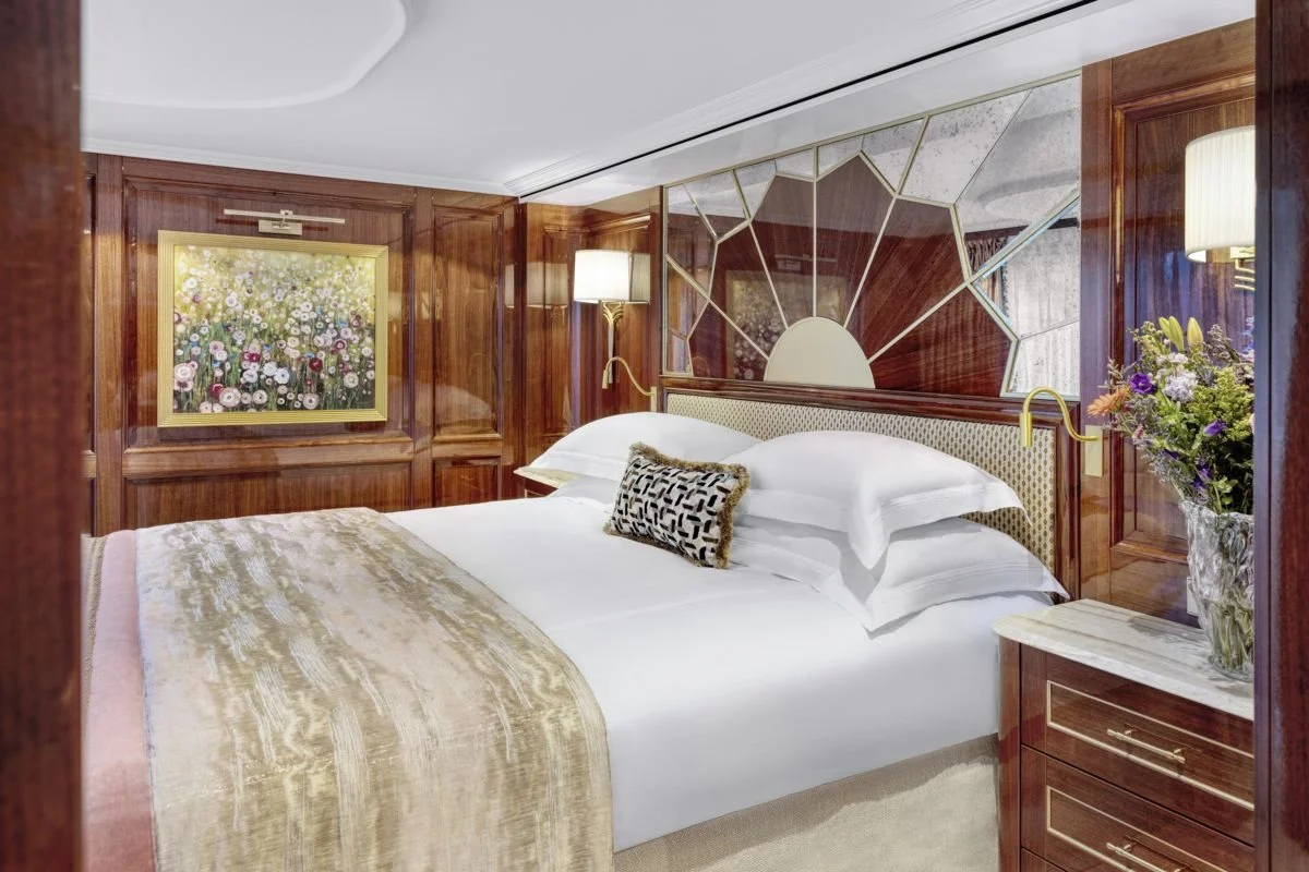 A luxurious hotel bedroom featuring a large bed with white linens, a velvet throw blanket, and decorative pillows. The wood-paneled walls include a floral painting, a geometric mirror, and a vase of flowers on a marble-top nightstand. There are wall-mounted lamps on either side of the bed.